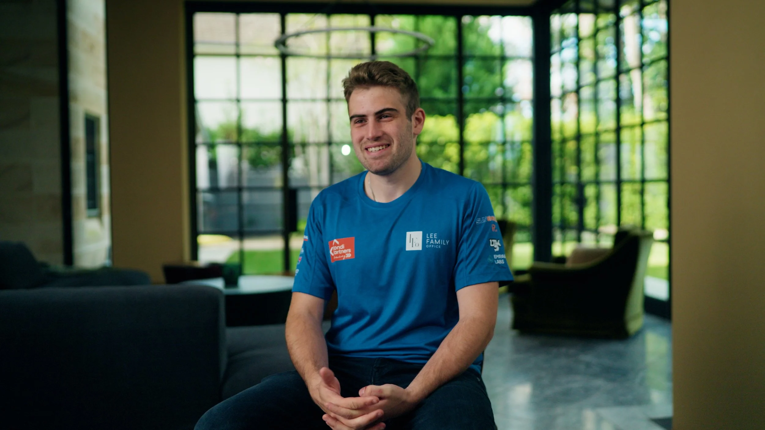 A young man with short brown hair and a light beard sitting indoors, smiling slightly, wearing a bright blue t-shirt with logos and text, in front of large windows showing a green outdoor area.