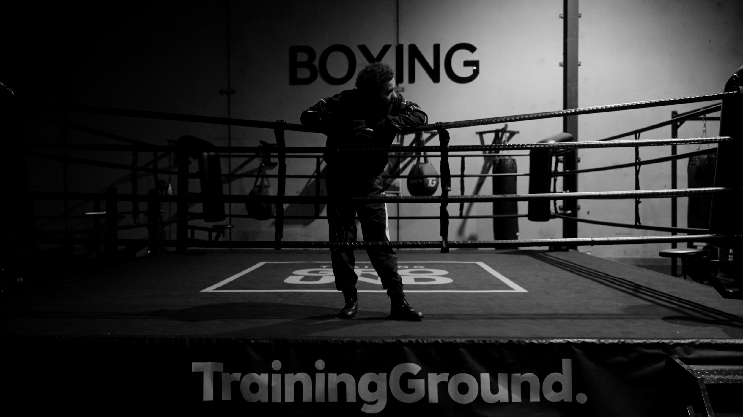 A man leaning on boxing ring ropes in a dimly lit gym, with large text on the wall behind him that reads 'BOXING' and part of a sign that says 'TrainingGround' in the foreground.