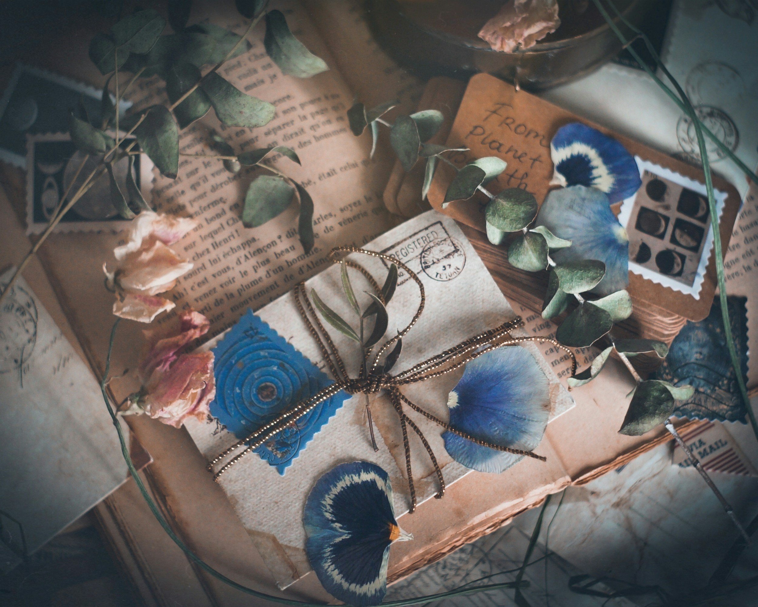 Close-up of a collection of vintage postage stamps, dried flowers, a handwritten note, and jewelry on a piece of paper or card. The note has French text and a postmark. The jewelry includes a gold chain and a ring.