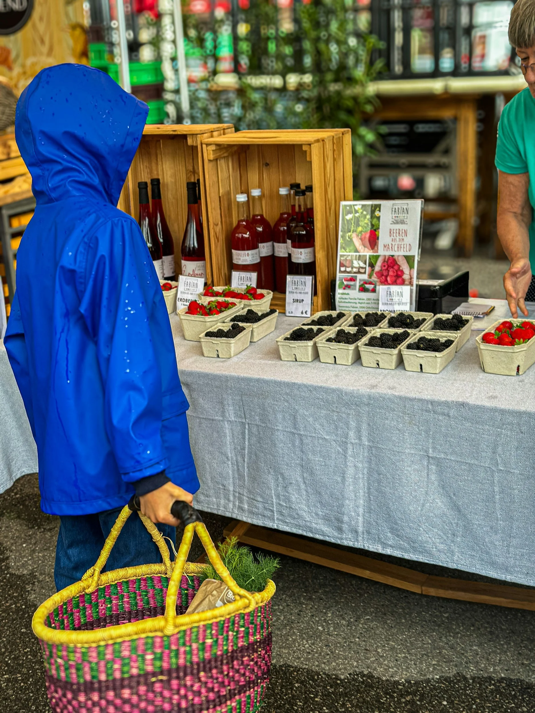A person in a blue raincoat carrying a colorful woven basket is looking at a market stall with jars of raspberry syrup, fresh strawberries, and blackberries. The stall has a sign advertising the produce and products.