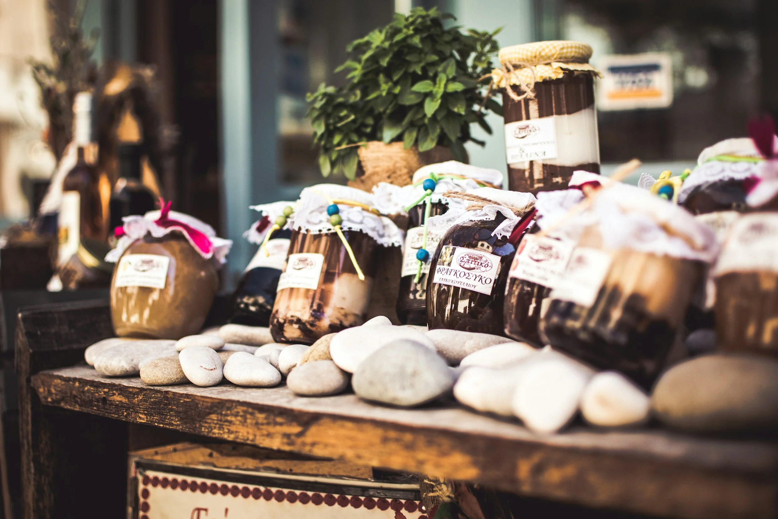 Photo of jars of homemade jam with fabric covers, a potted plant, and white stones on a wooden display table outside.