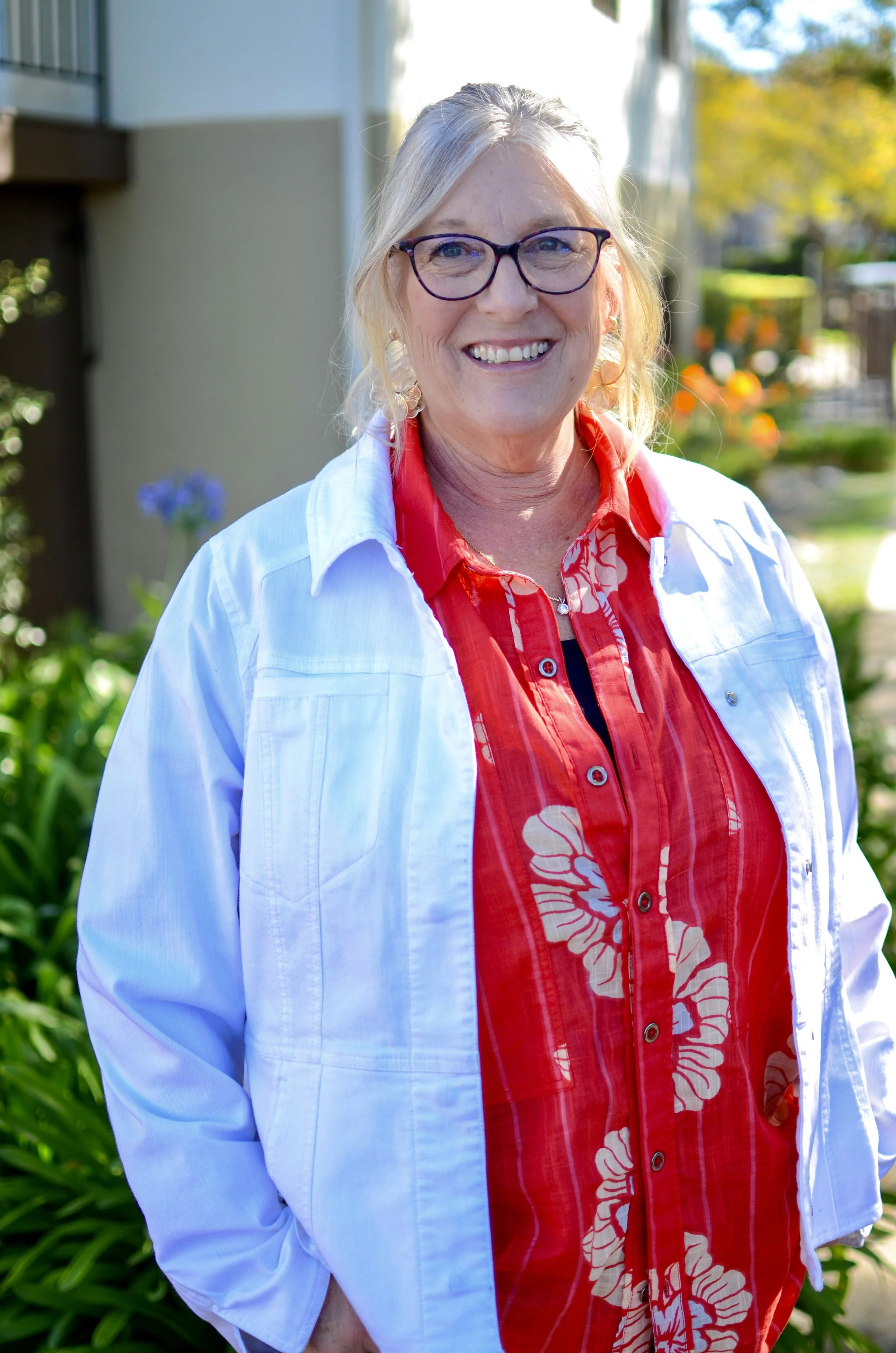 A smiling woman with glasses, wearing a red and white patterned shirt and a white jacket, standing outdoors with a background of trees and plants.
