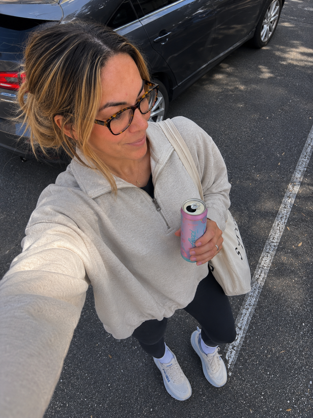 Haus of Whit Brand Photoshoot 2026. Whit Radacina taking a selfie in a parking lot, holding a can of sparkling water. She is wearing a beige fleece jacket, black pants, and white sneakers.