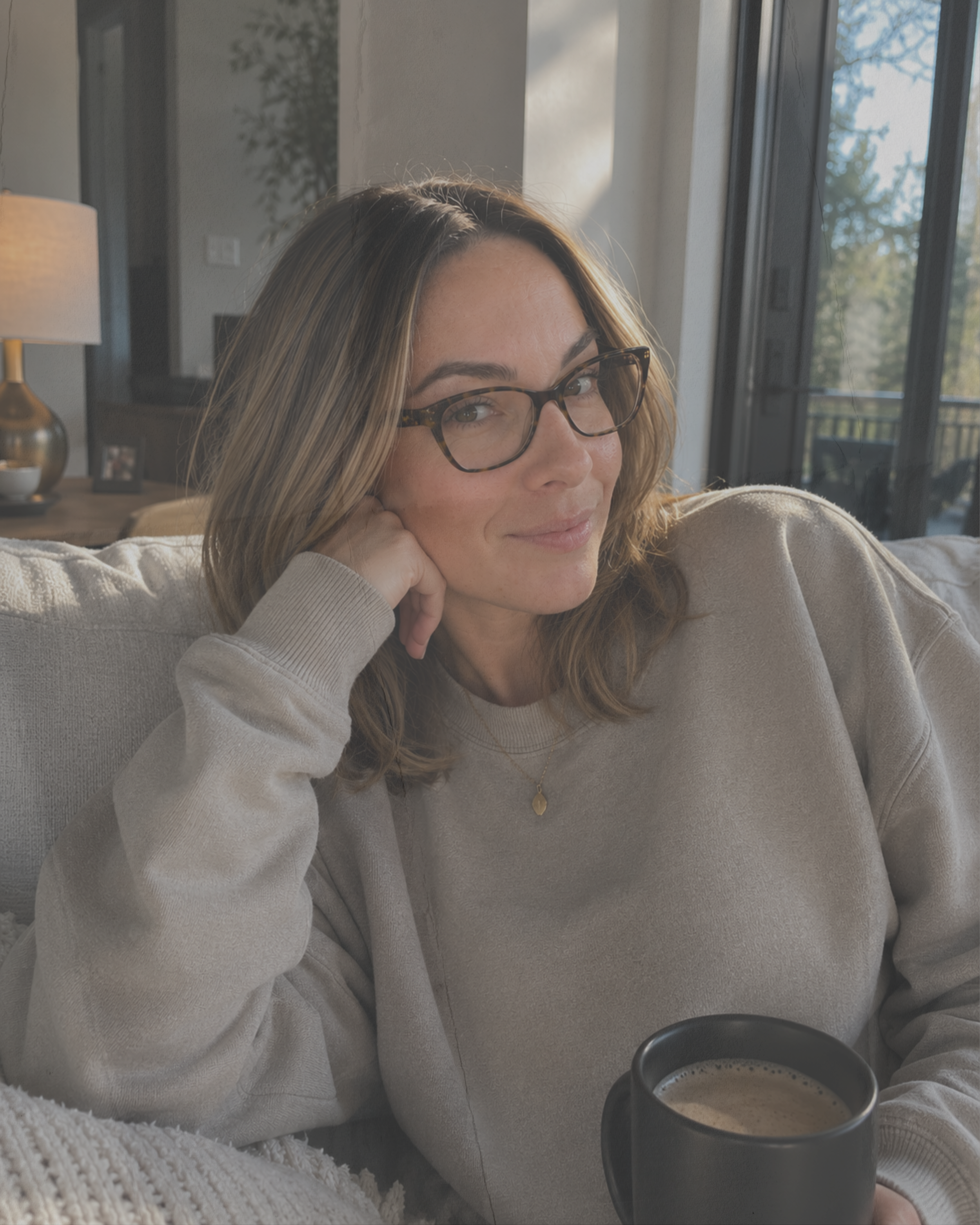 Haus of Whit Brand Photoshoot 2026. Whitney Radacina sits on a couch, resting her chin on her hand, in a cozy living room with sunlight streaming through large windows, holding a mug of coffee or tea.