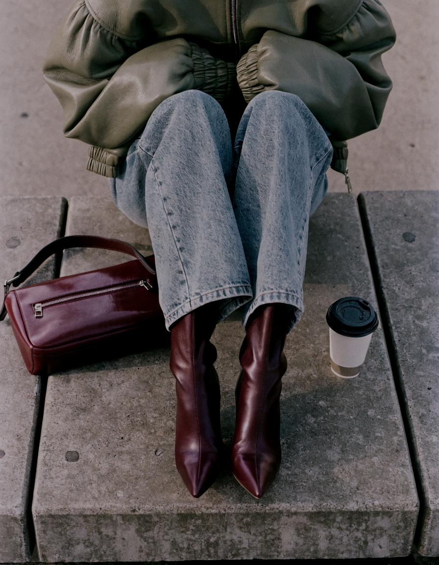 Haus of Whit, Whitney Radacina Brand Photoshoot 2026.  A woman sitting on a concrete bench, wearing a green jacket, light blue jeans, and burgundy high-heeled boots, with a burgundy handbag and a to-go coffee cup next to her.