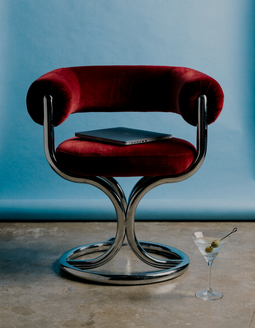 Haus of Whit, Whitney Radacina Brand Photoshoot 2026. A modern red velvet chair with a chrome frame, a closed laptop on the seat, and a martini glass with an olive on a cocktail pick placed on the floor next to it, against a light blue wall.
