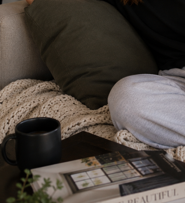 Haus of Whit Brand Photoshoot 2026. Close-up of a cozy living room corner with a beige chunky knit blanket, a black mug, a magazine on a dark wooden table, and Whit Radacina sitting on a gray couch wearing gray sweatpants and a black shirt.