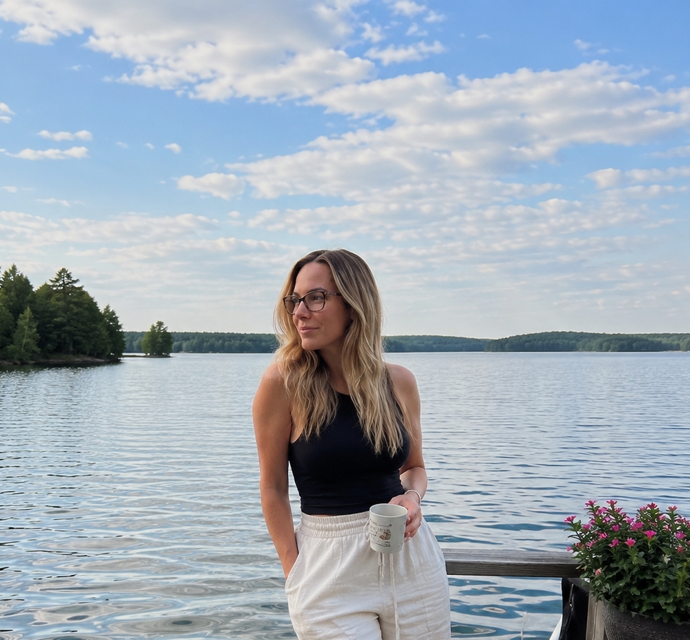 Haus of Whit Brand Photoshoot 2026. Whitney Radacina, wearing a black tank top and white pants, holding a mug, standing near a lake with a tree-lined shore, blue sky, and scattered clouds.