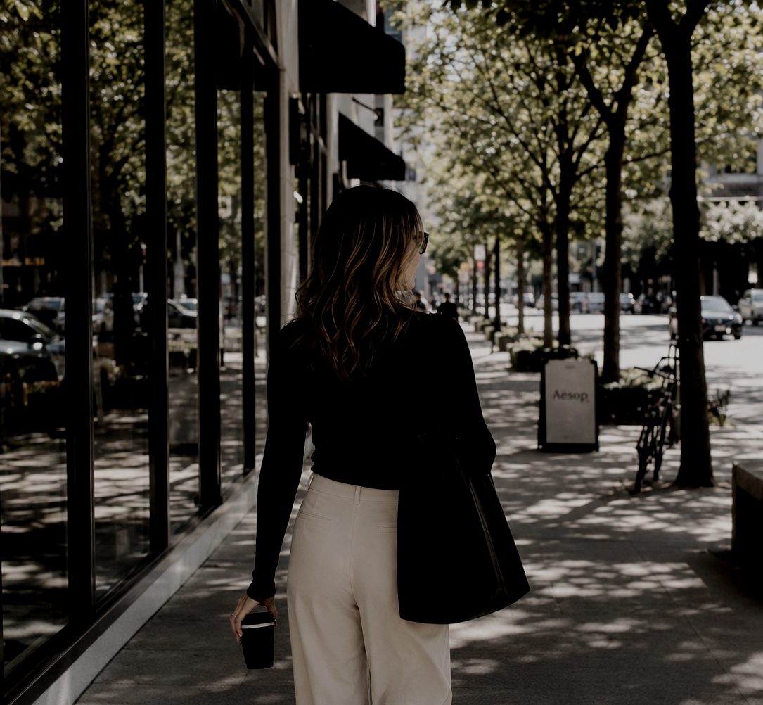 Haus of Whit Brand Photoshoot 2026. Whitney Radacina dressed in a black top and light-colored pants, walking along a tree-lined city sidewalk while holding a coffee cup, with shops and cars in the background.