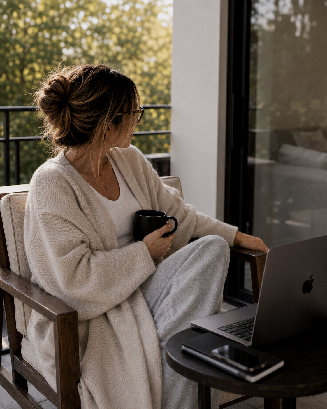 Haus of Whit Brand Photoshoot 2026. Whitney Radacina with a messy bun hairstyle sitting on a balcony, holding a black coffee mug, with a laptop and phone on a small round table beside her, looking outside.