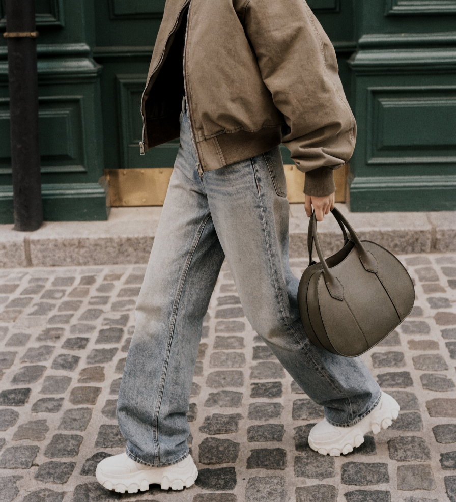 Haus of Whit, Whitney Radacina Brand Photoshoot 2026.  Person walking on cobblestone street carrying a gray bag, wearing a brown jacket and loose jeans, with white chunky sneakers.