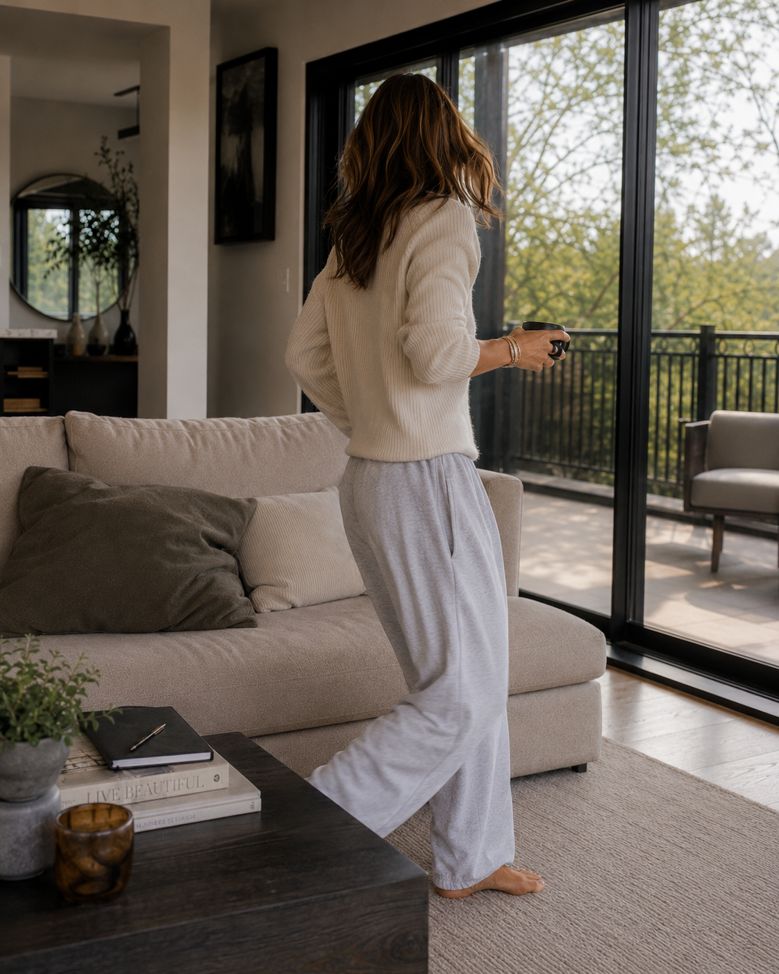 Haus of Whit Brand Photoshoot 2026. Whitney Radacina in a cozy living room, wearing a white sweater and light gray pajama pants, holding a mug, standing near a glass sliding door overlooking an outdoor balcony with trees.