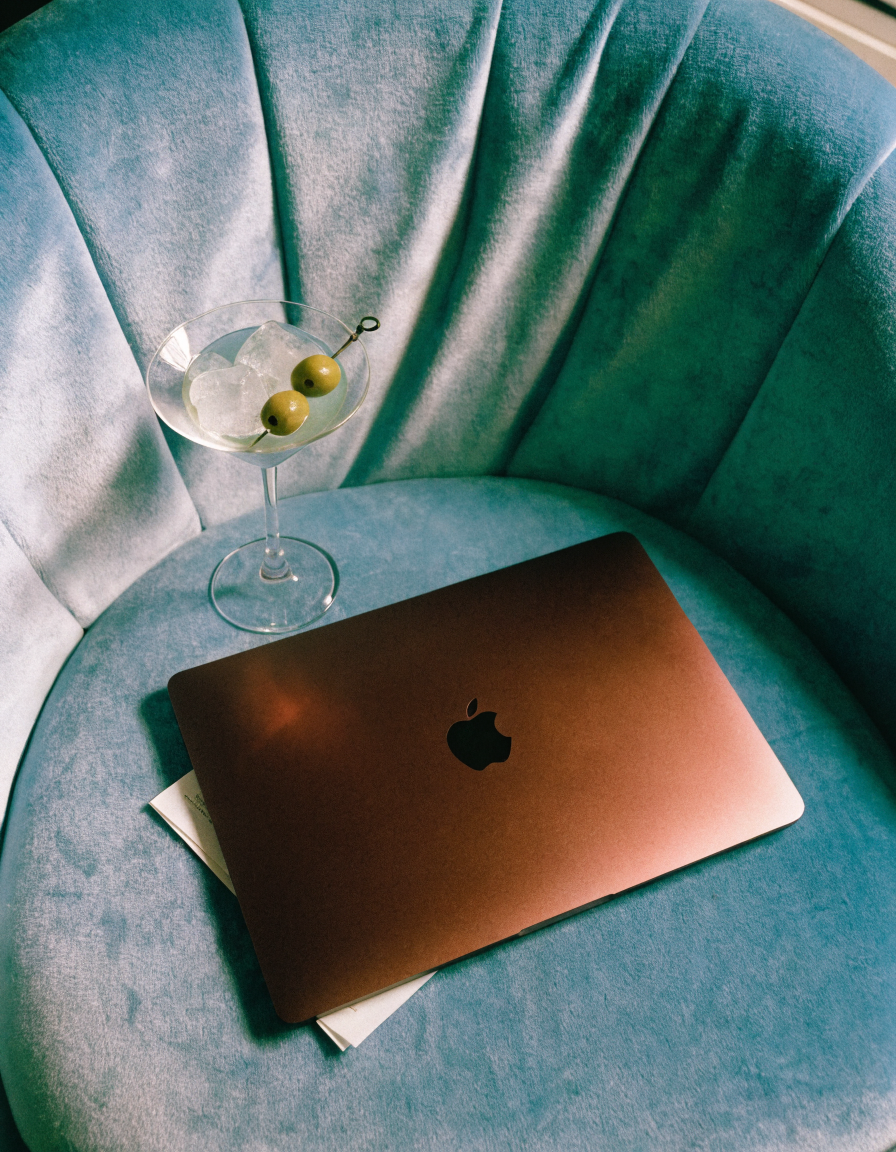 Haus of Whit, Whitney Radacina Brand Photoshoot 2026. A closed rose gold MacBook on a blue velvet curved sofa. A martini glass filled with ice and garnished with two green olives rests on the sofa beside the MacBook.