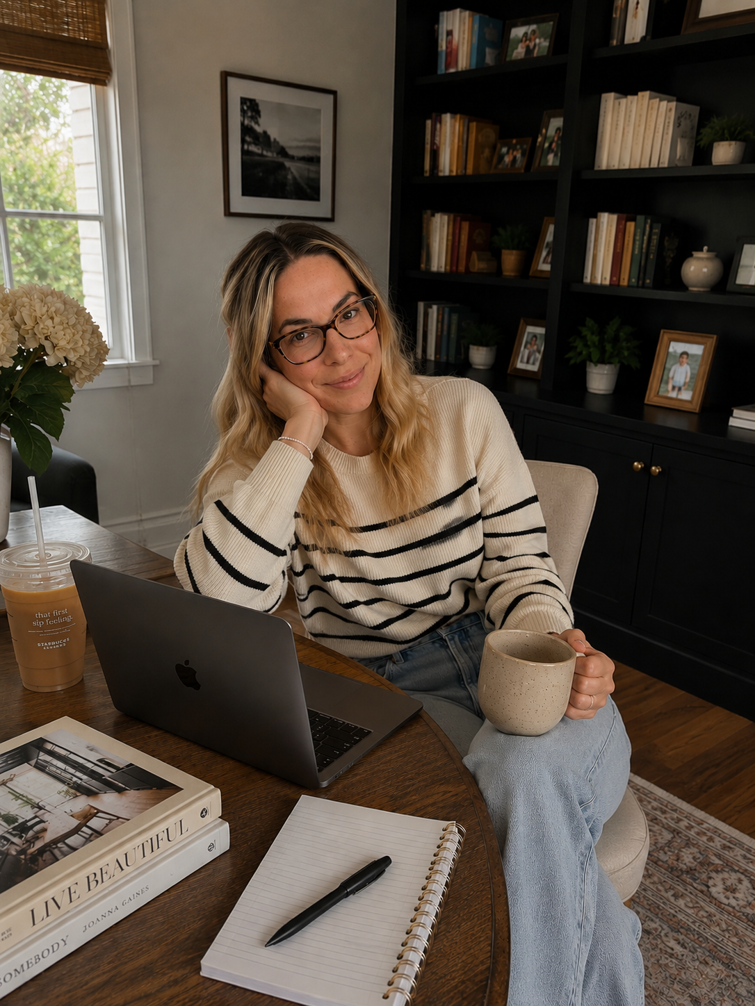 A woman with blonde hair and glasses sits at a wooden table with a laptop, a notebook, a pen, a book titled 'Live Beautiful,' and a coffee cup. She holds another coffee cup in her hand and looks content. The background features a bookshelf with books and framed photos, and a window with a view outside.