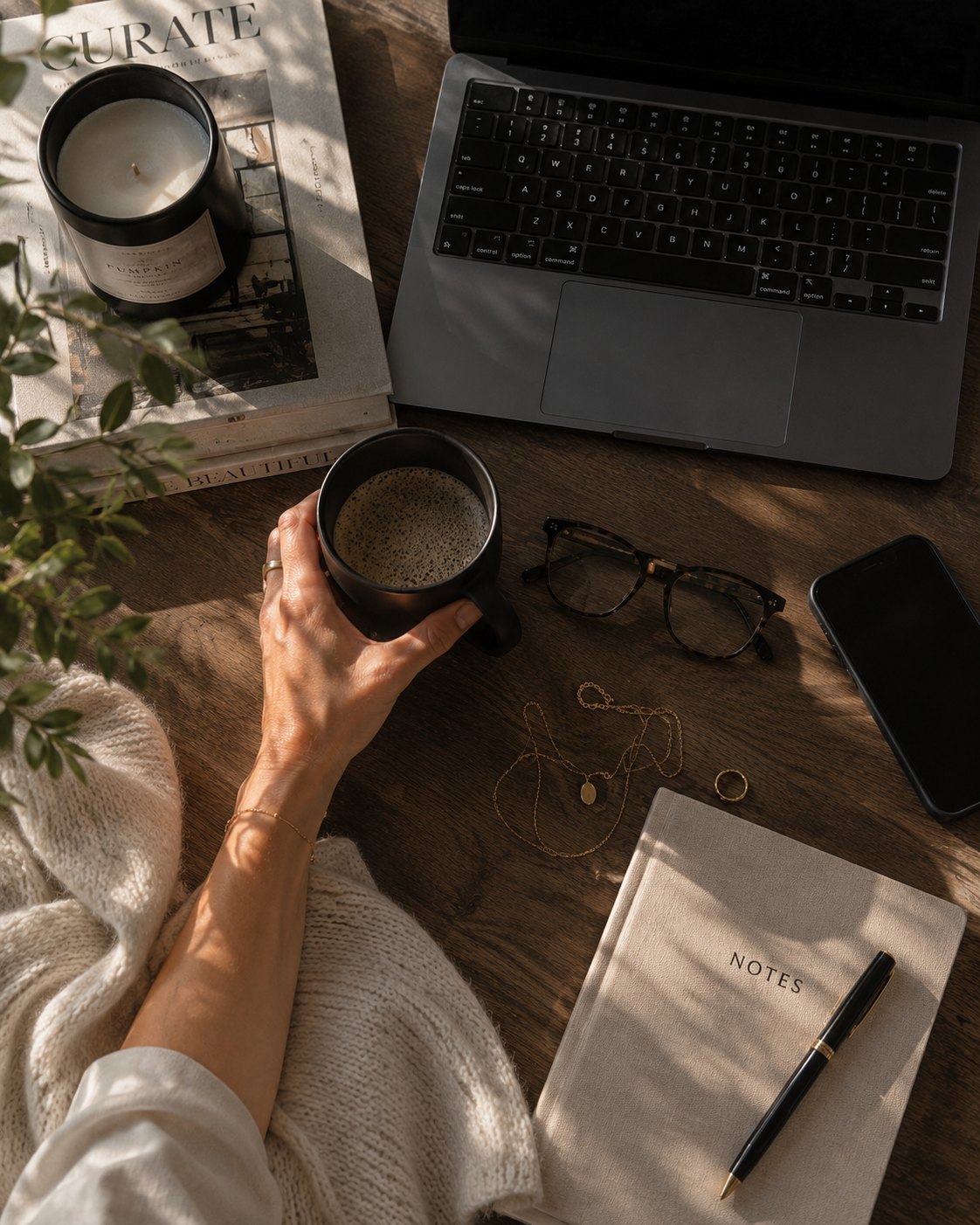 Haus of Whit Brand Photoshoot 2026. A workspace flatlay with Whitney Radacina holding a black mug of coffee, a laptop, a smartphone, eyeglasses, gold jewelry, a notebook, pen, candle, and a plant, bathed in natural light.