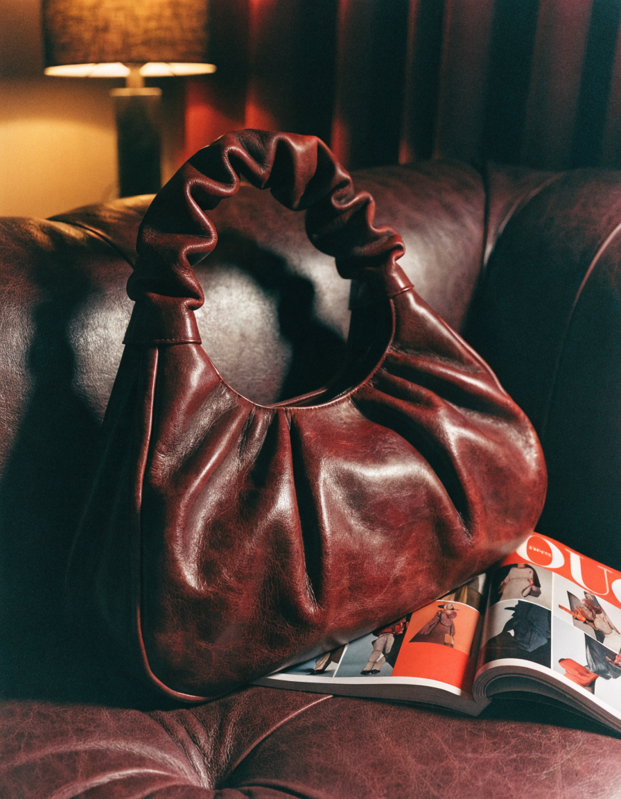 Haus of Whit, Whitney Radacina Brand Photoshoot 2026.  A dark red leather handbag resting on a leather couch with an open magazine beside it.
