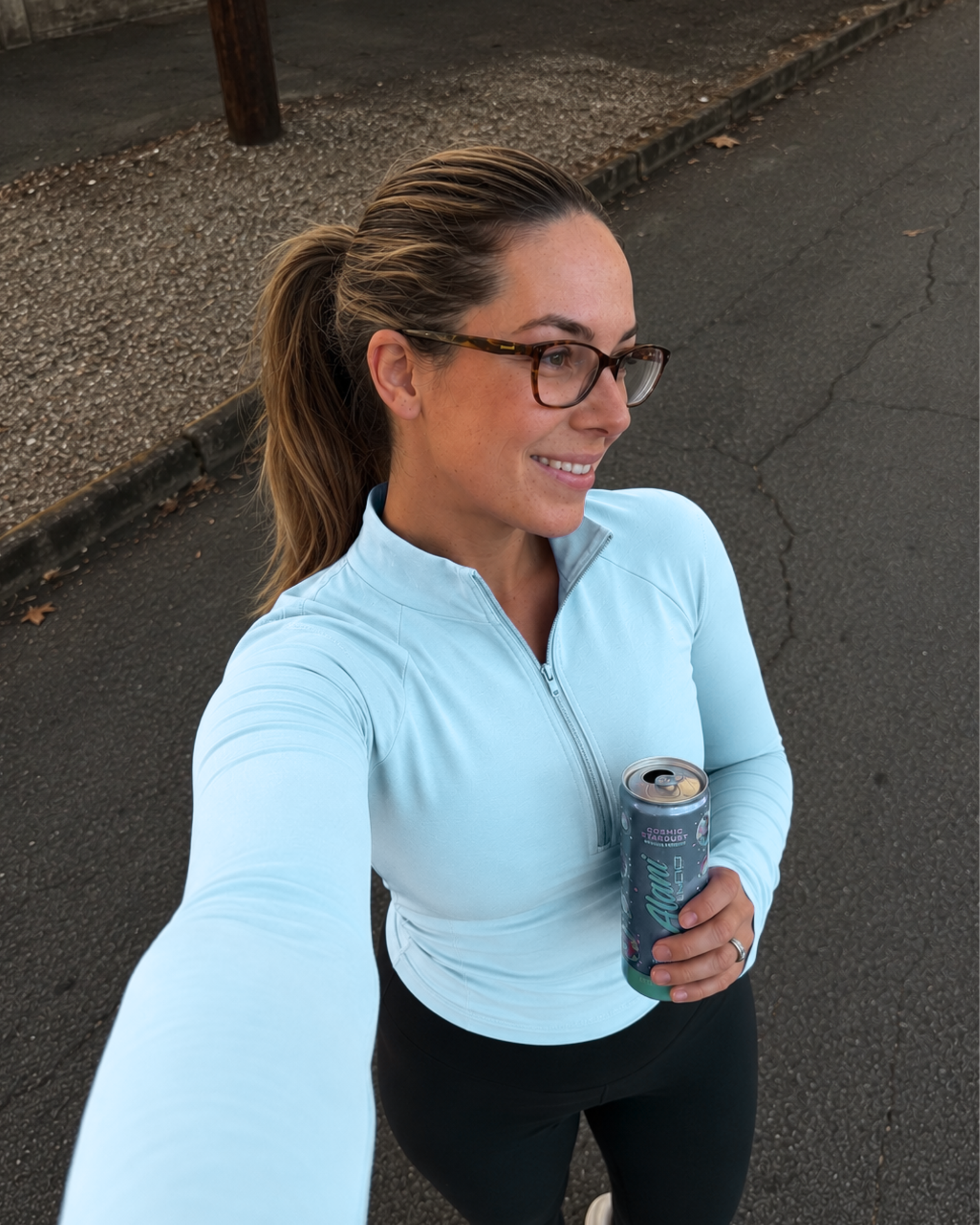 Haus of Whit Brand Photoshoot 2026. Whitney Radacina smiling, wearing a light blue jacket, holding a can of LaCroix sparkling water, standing on a paved street at night with a curb and some fallen leaves visible.