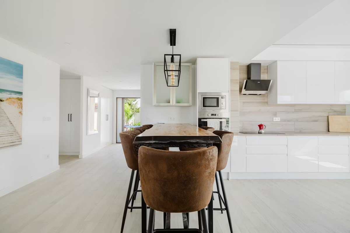 Modern open-concept kitchen with a wooden dining table, brown chairs, white cabinets, built-in microwave, and a black range hood.
