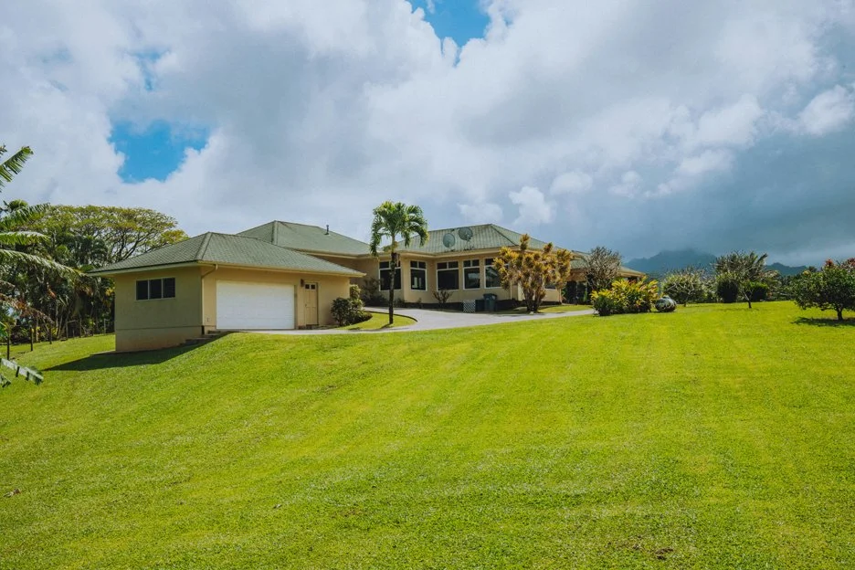 Large house with a green roof and a two-car garage, surrounded by a lush green lawn, palm trees, and other vegetation, under a partly cloudy sky.