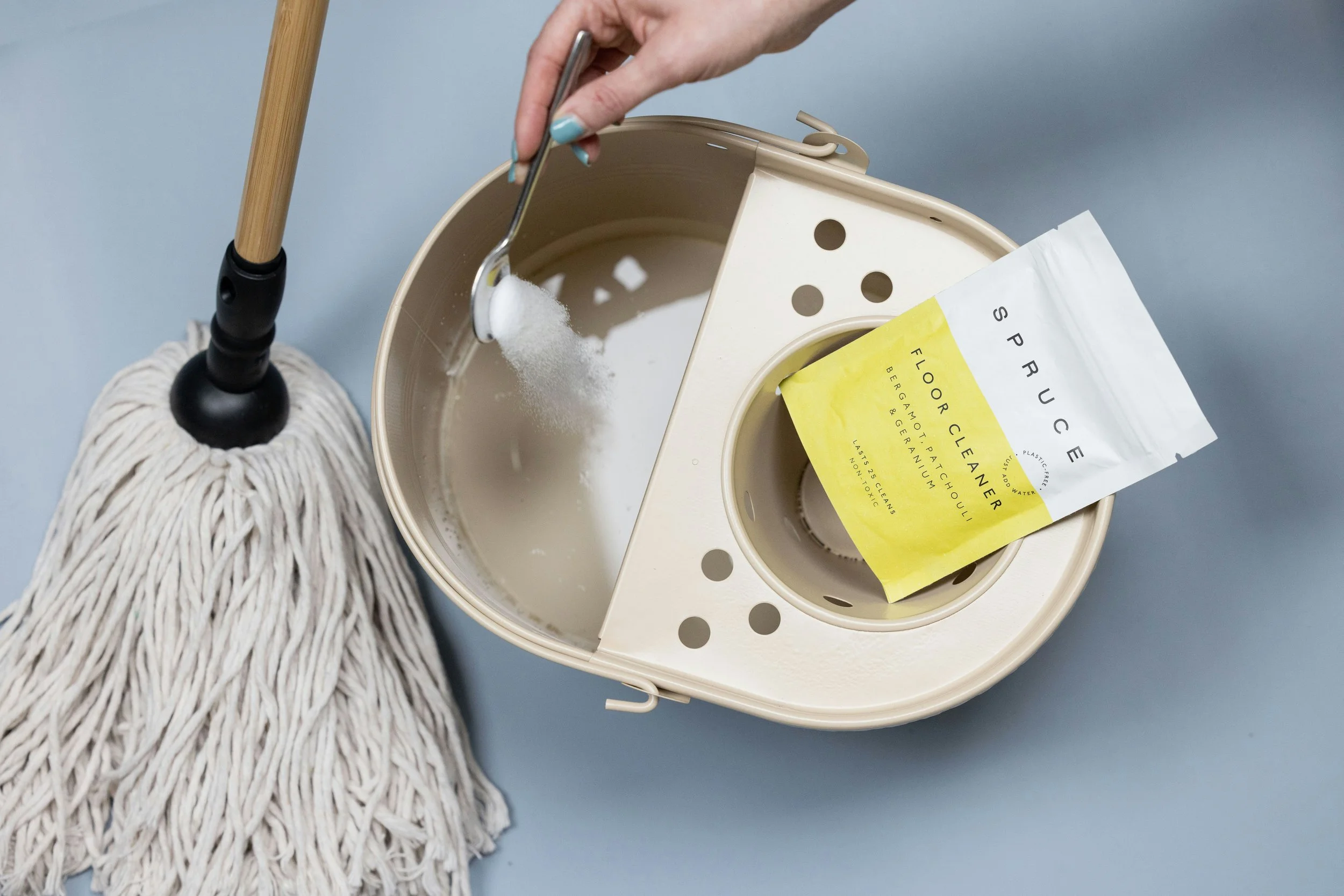 Hand holding a spoonful of white powder over a beige, round, vintage-style trash can with a paper bag labeled 'Floor Cleaner' inside. A mop with a wooden handle and a fluffy, twisted cotton mop head leans against the trash can. The background is a plain, light blue wall.