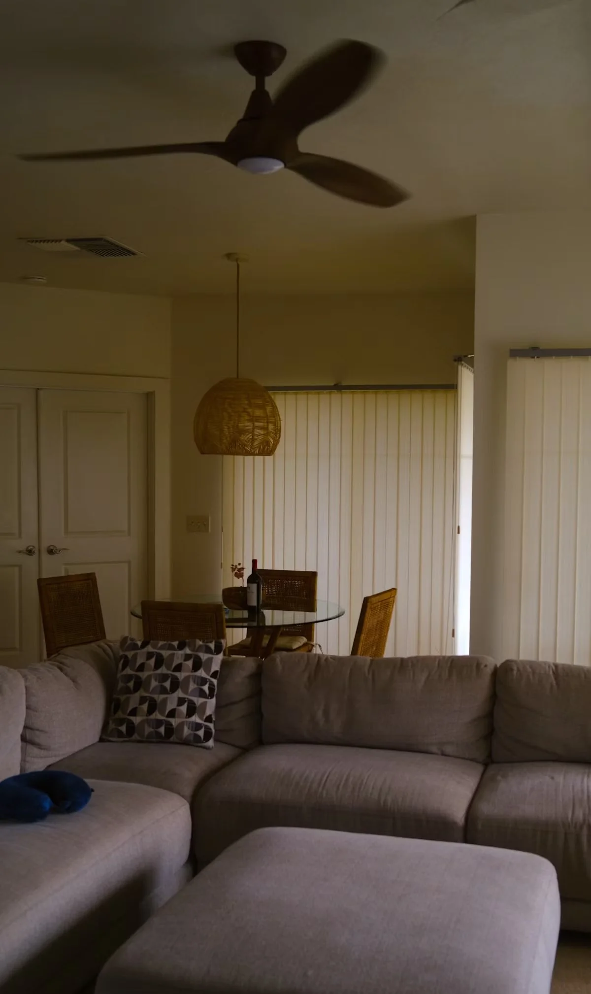 Living room with a beige sectional sofa, a patterned pillow, and a matching ottoman in the foreground. In the background, there's a dining area with a glass table, rattan chairs, a bottle of wine, and a hanging wicker light. A ceiling fan with wooden blades is mounted on the ceiling.