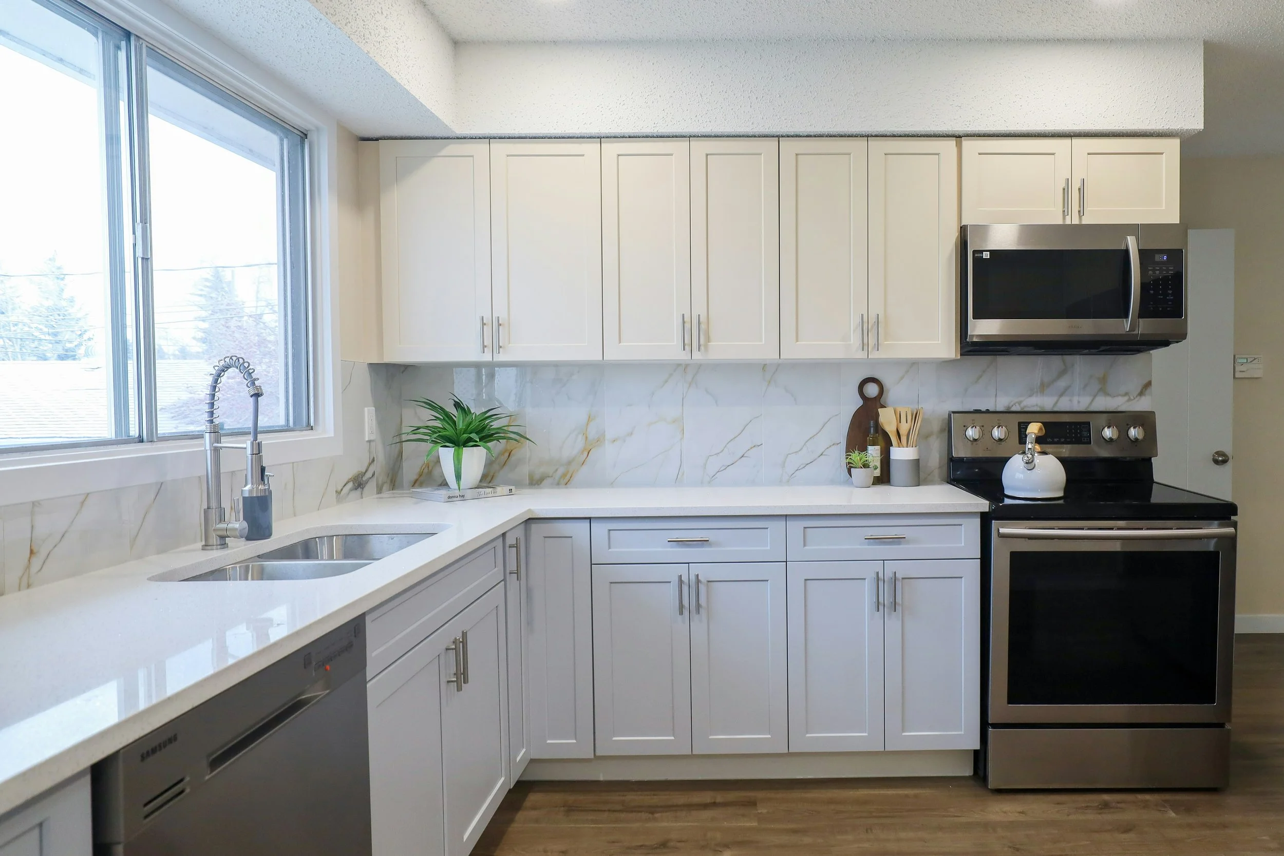 Modern kitchen with white cabinets, marble backsplash, stainless steel appliances including a microwave and oven, a window with a view outside, and small potted plants on the counter.