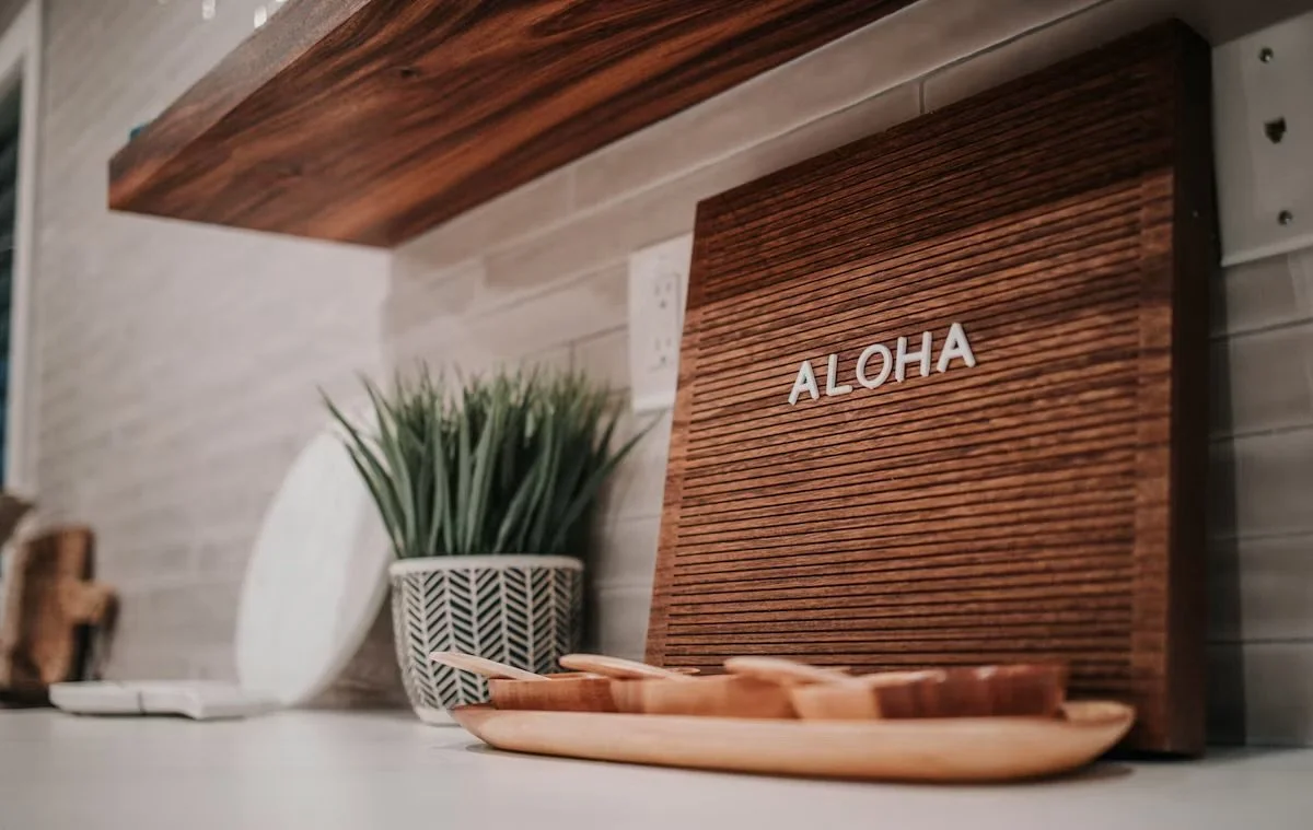 Decorative kitchen shelf with a wooden sign reading 'Aloha', a potted plant, and a wooden tray, against a tiled wall.