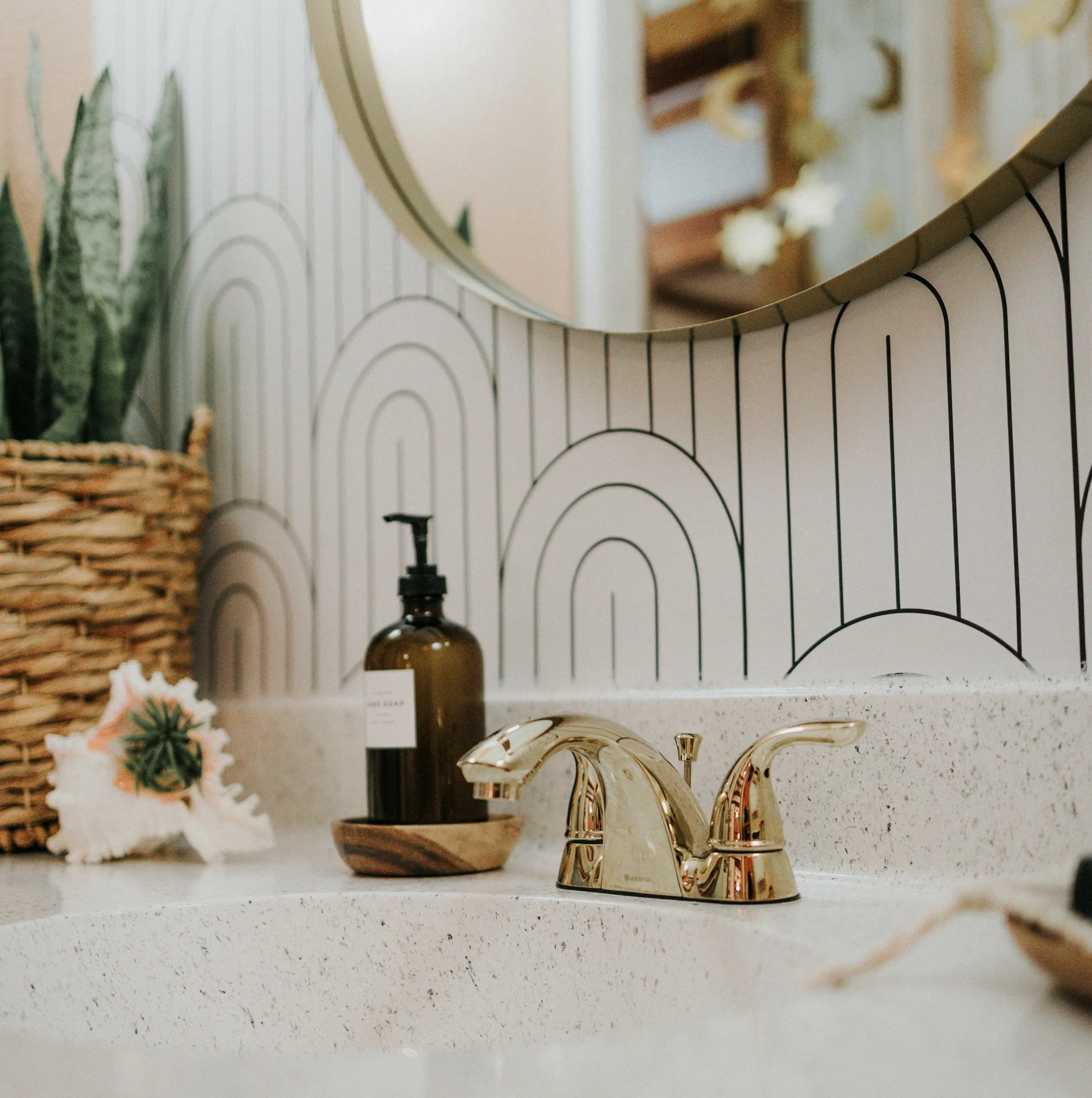 Close-up of a shiny gold faucet with a modern curved design on a light-colored speckled countertop. In the background, a small brown vase and decorative items are visible, along with a striped and patterned backsplash.