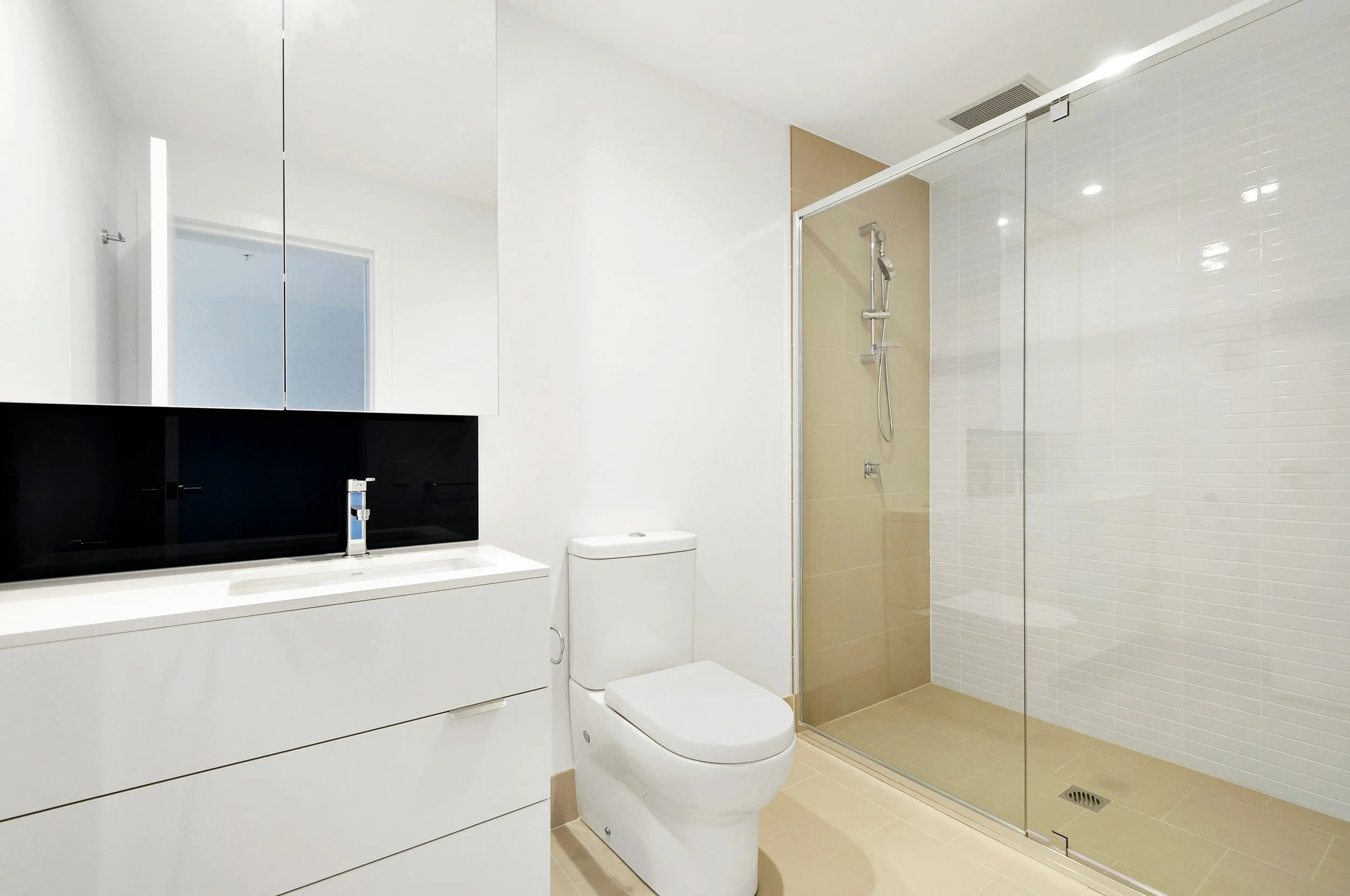 Modern bathroom with white walls, a glass-enclosed shower with beige tiles, a white toilet, and a white vanity with a black countertop and a mirror above.