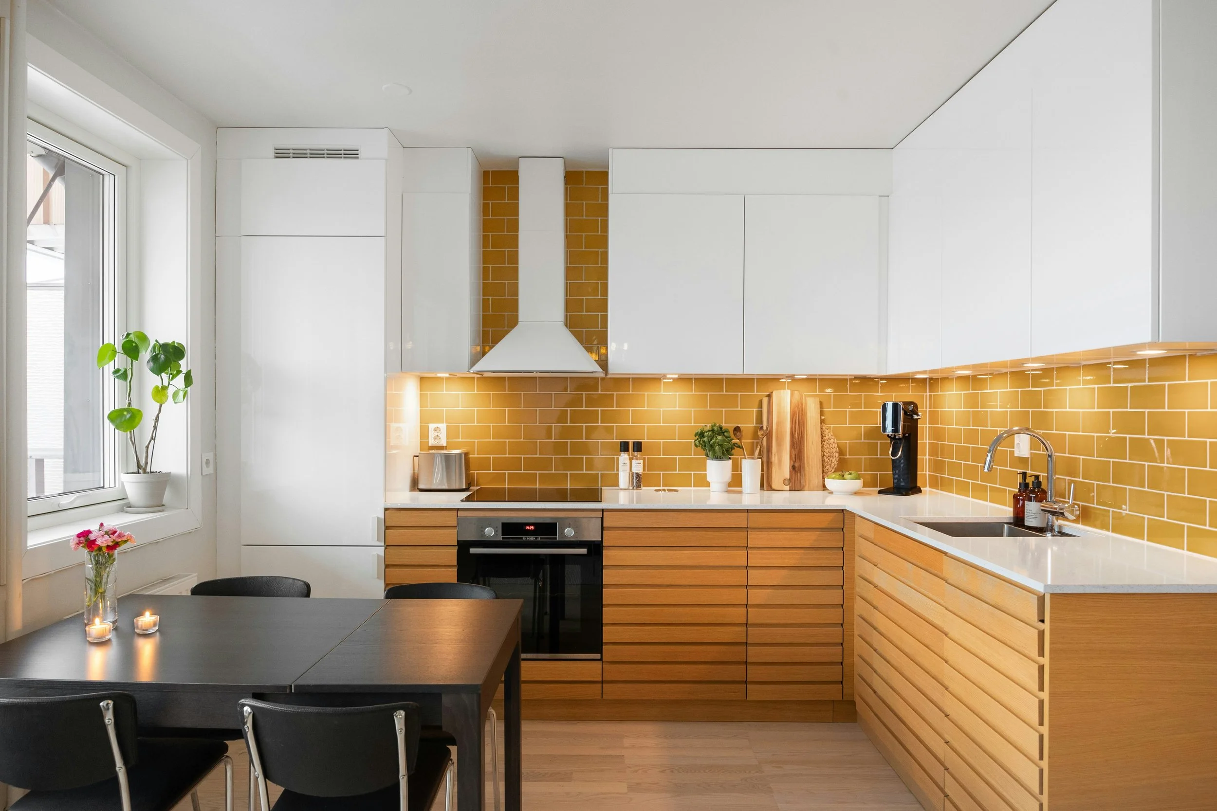 Modern kitchen with white upper cabinets, wooden lower cabinets, yellow subway tile backsplash, countertop with small potted plant, chopping board, coffee machine, and sink. A window with a potted plant and candles on the table in the foreground.