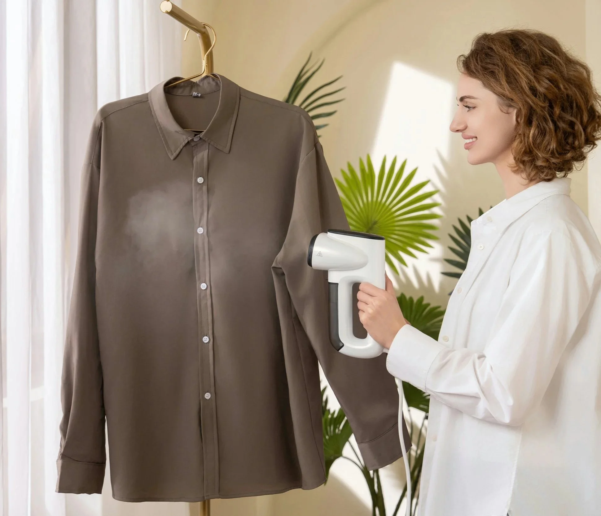 A woman with curly hair using a handheld scanner on a brown long-sleeve shirt hung on a hanger, in a room with natural light and green indoor plants.