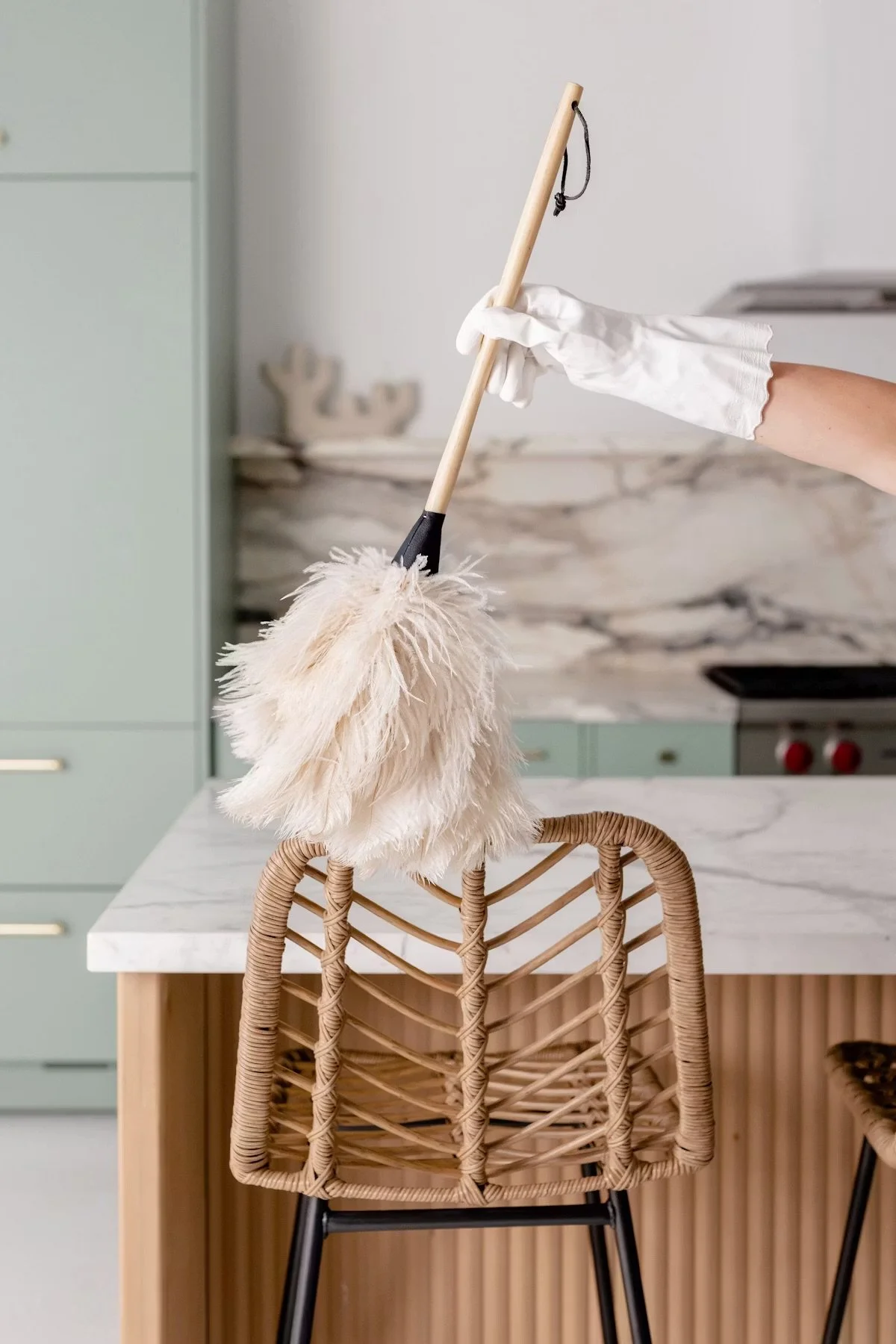 Hand wearing a white glove holding a cleaning duster over a woven chair in a modern kitchen.