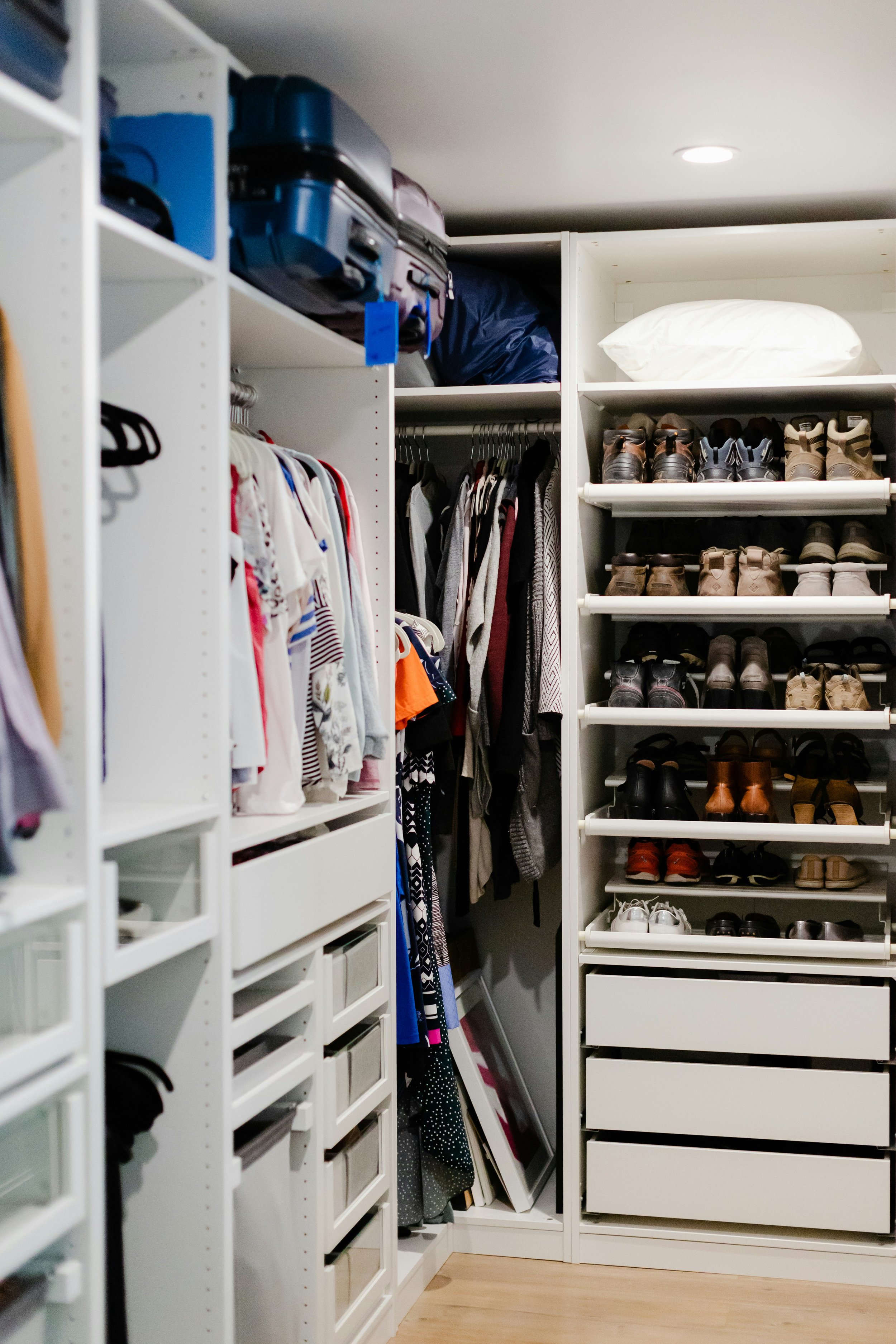 A walk-in closet with white shelving units filled with clothes, shoes, and suitcases, with a wooden floor and a ceiling light.