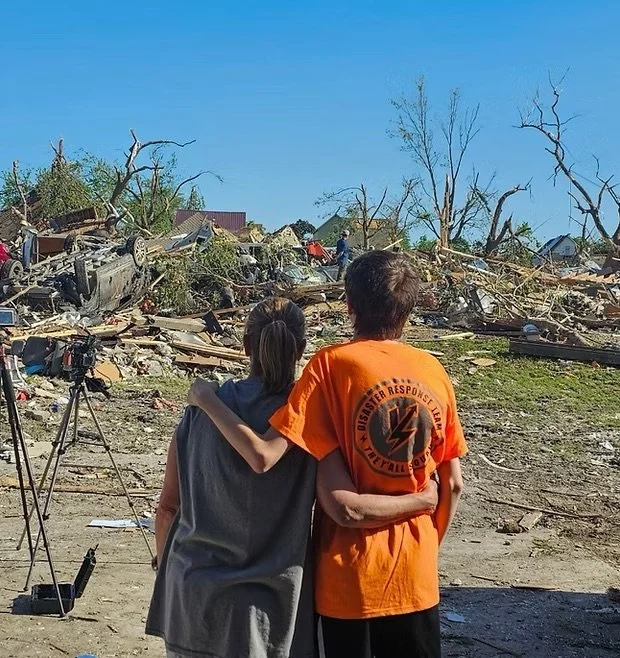 Two people embrace amid tornado damage, overturned vehicles, and fallen trees
