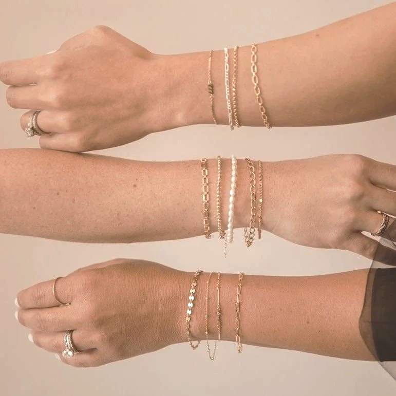 Three arms extended and overlapping, each wearing multiple gold and pearl bracelets and rings from Avec Toi Permanent Jewelry.