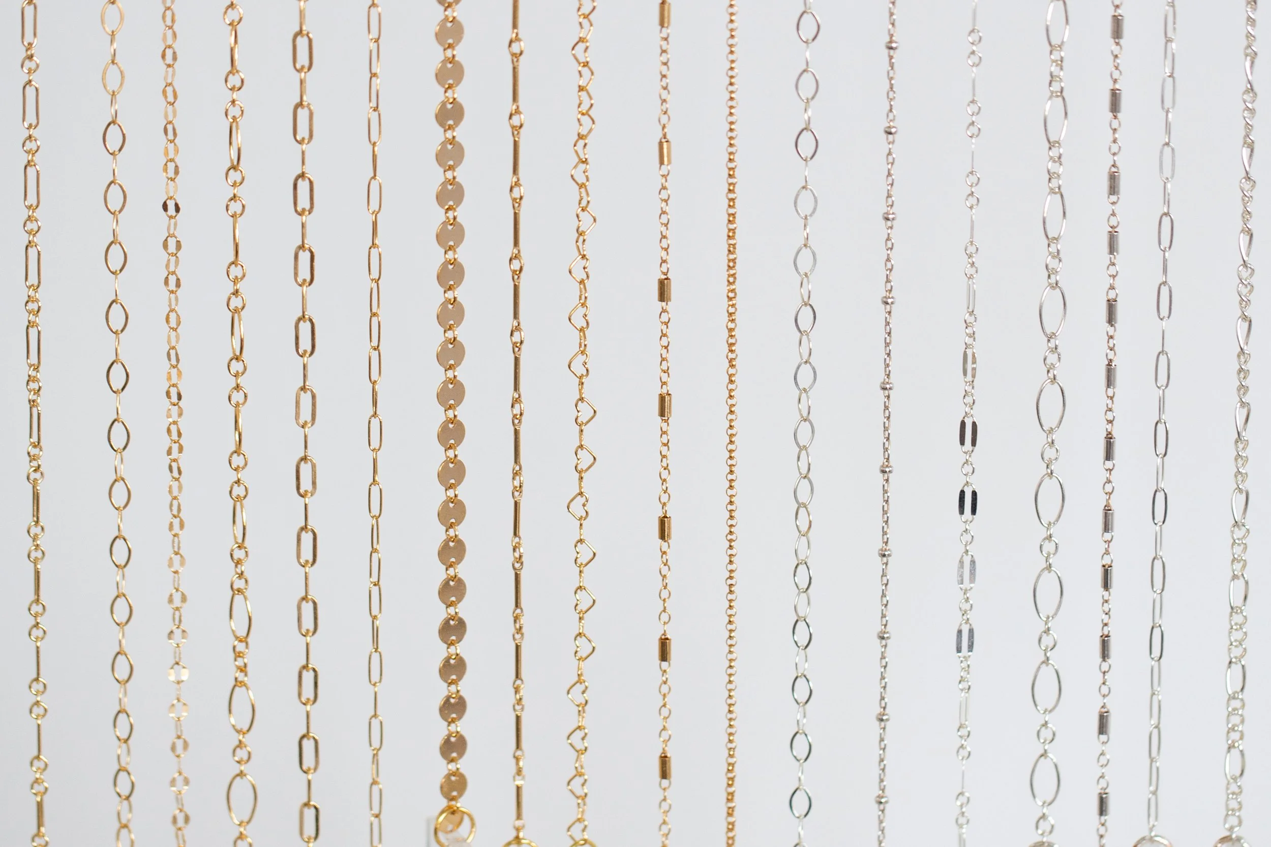Multiple silver and gold chains hanging vertically against a light background from Avec Toi Permanent Jewelry.
