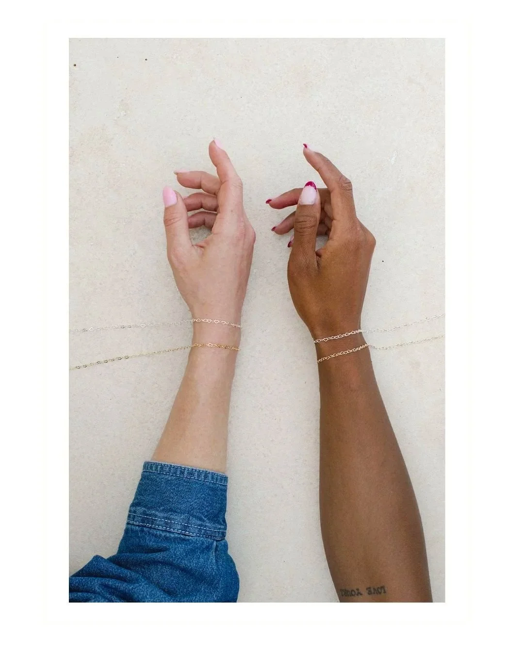 Two hands with bracelets on wrists, one with light skin and the other with dark skin, reaching towards each other, on a beige surface.