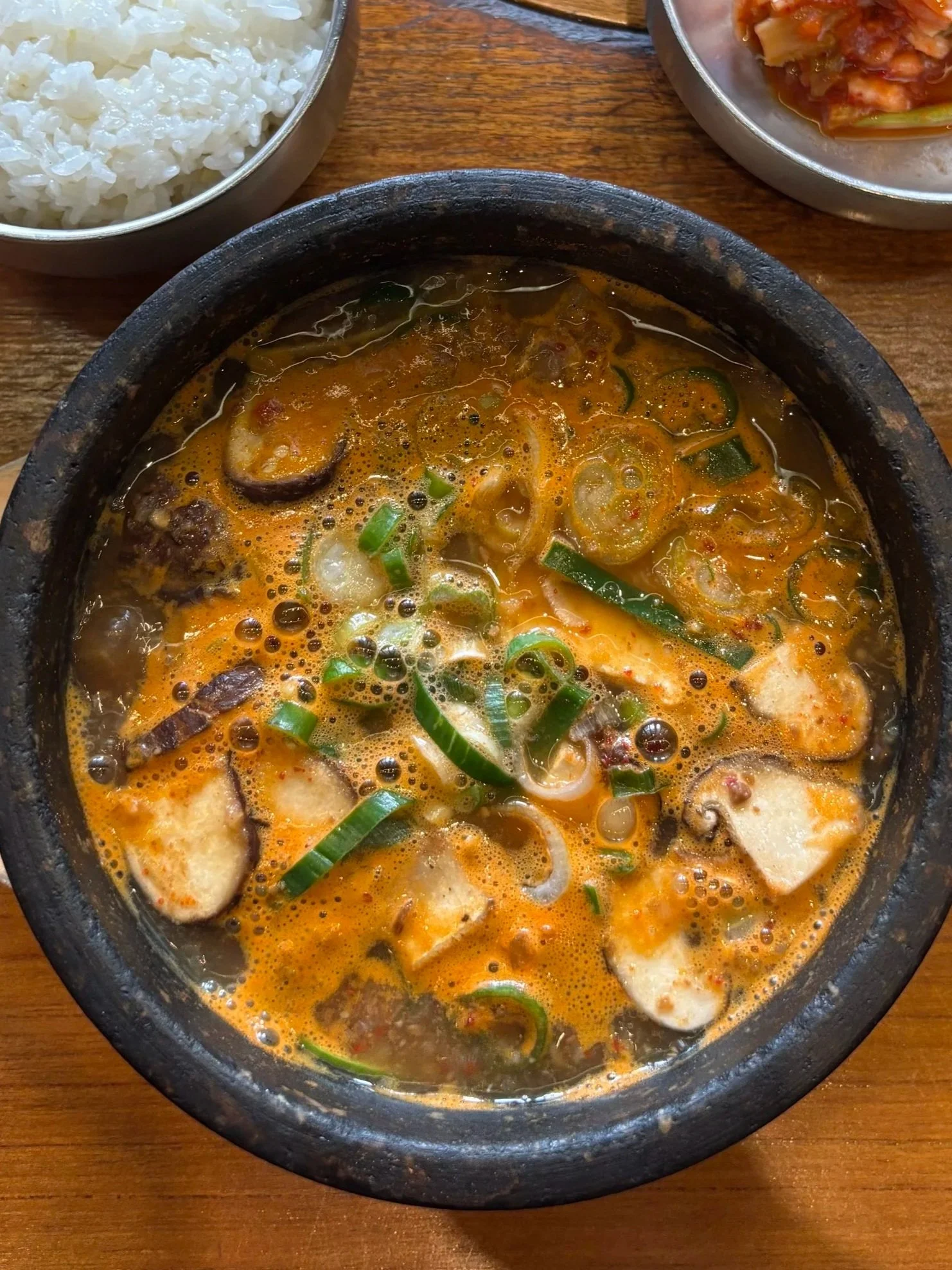 A bowl of spicy Korean soup with green onions, mushrooms, and fish cake, surrounded by bowls of white rice and kimchi on a wooden table.