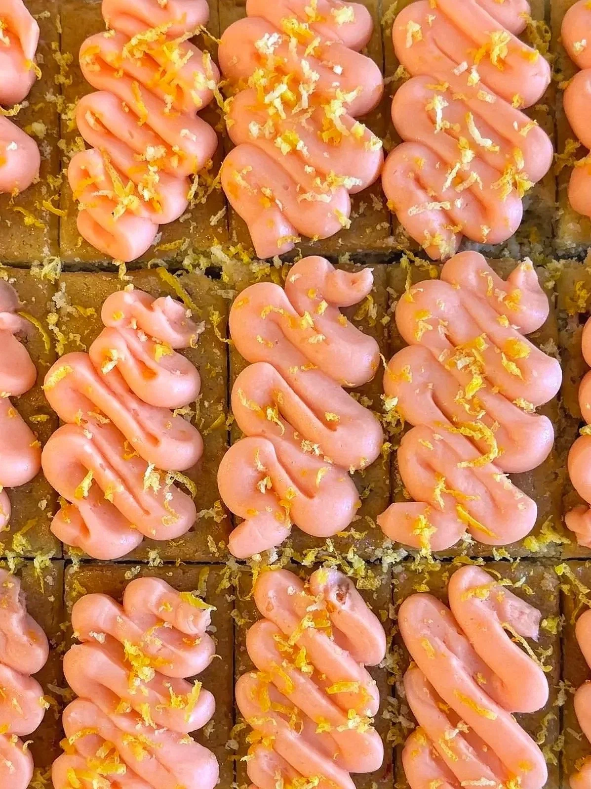Close-up of lemon bars of topped with pink frosting swirled in a zigzag pattern and garnished with yellow zest, arranged in a grid.