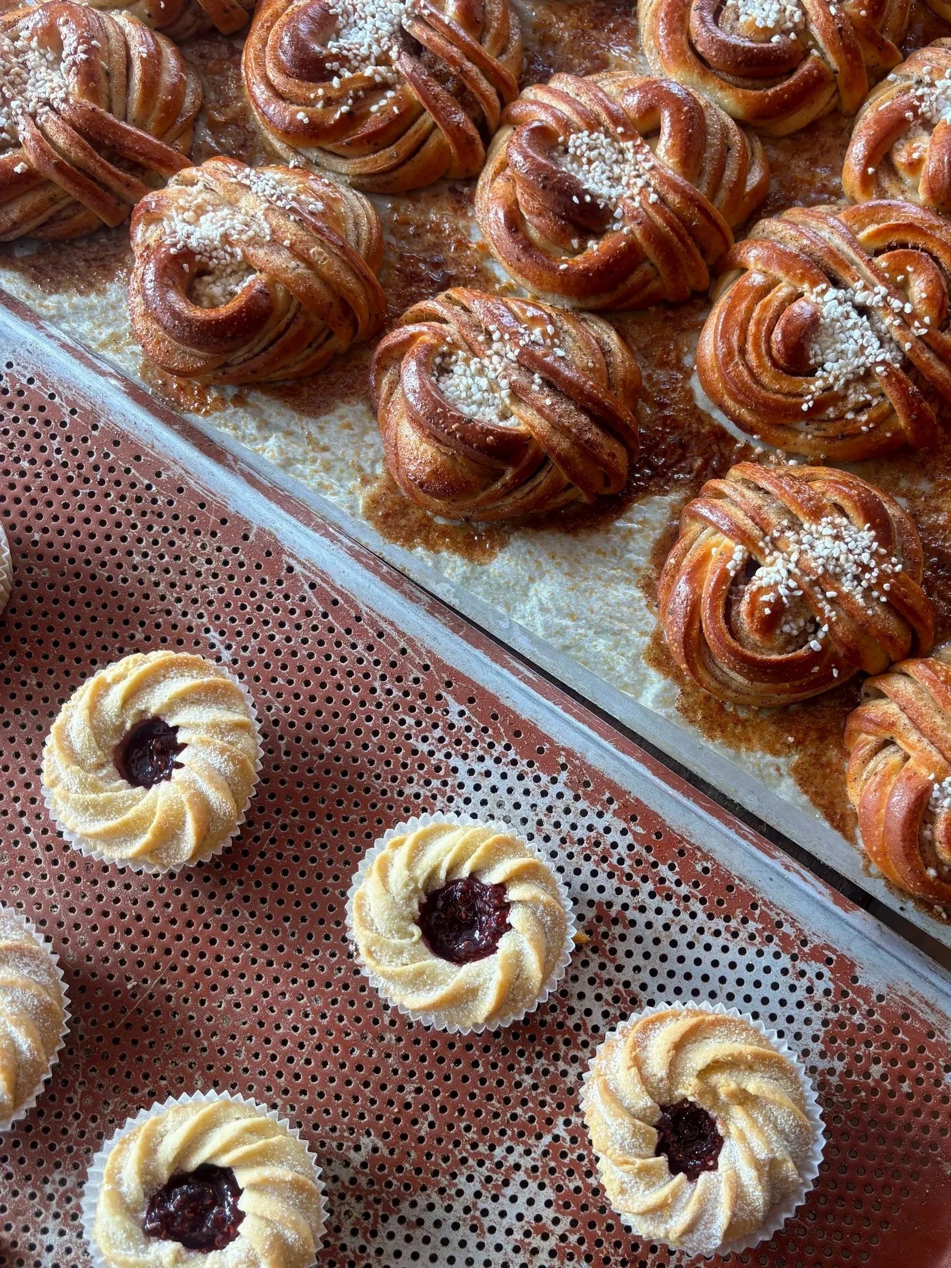 Close-up of freshly baked Swedish cinnamon rolls, topped with pearl sugar, in a baking tray, with some raspberry jam-filled cookies on a perforated surface.