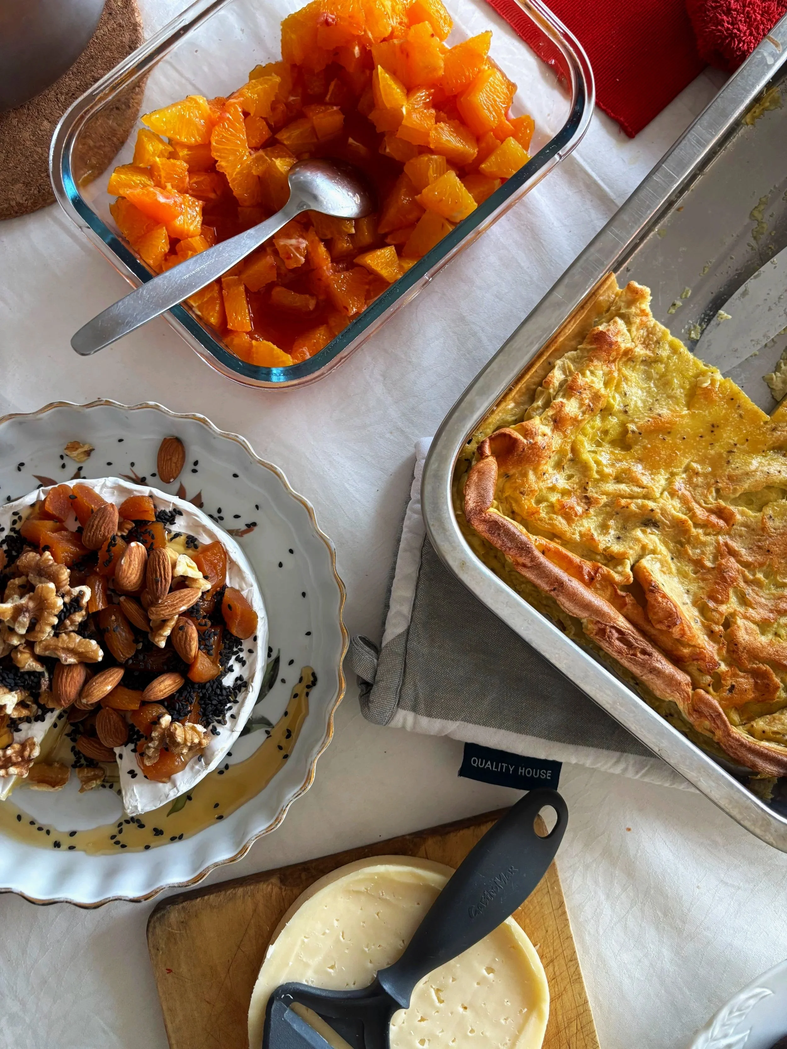 A dish of orange fruit chunks in a glass container, a cheese wheel topped with nuts and dried fruit, and a partially sliced wheel of cheese on a wooden cutting board.