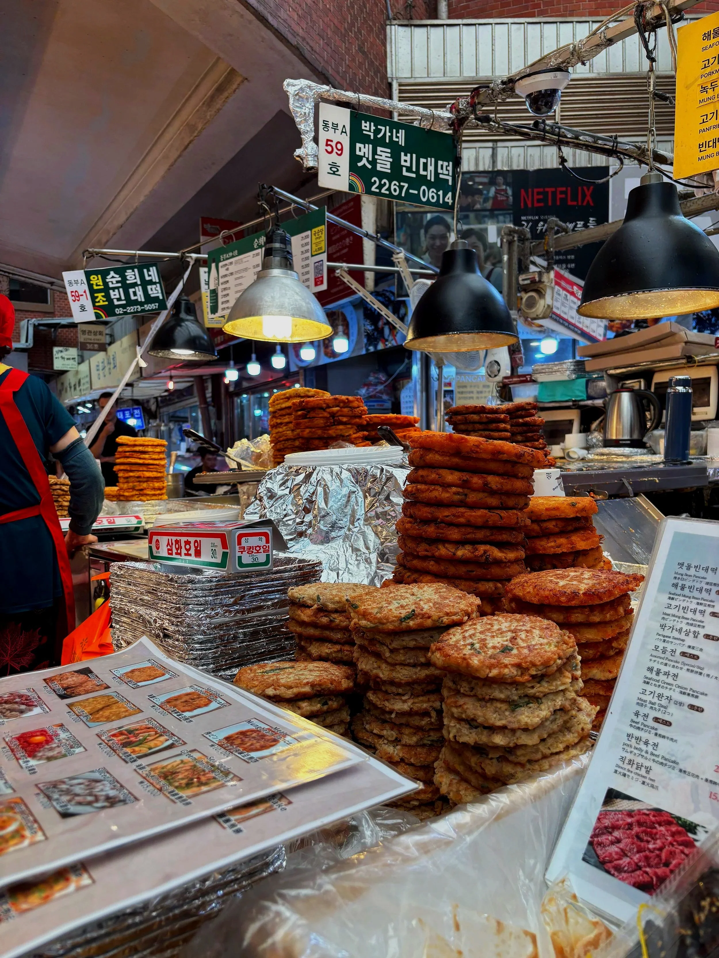 Stacked various types of Korean pancakes on a food stall table with a menu and signs in Korean.