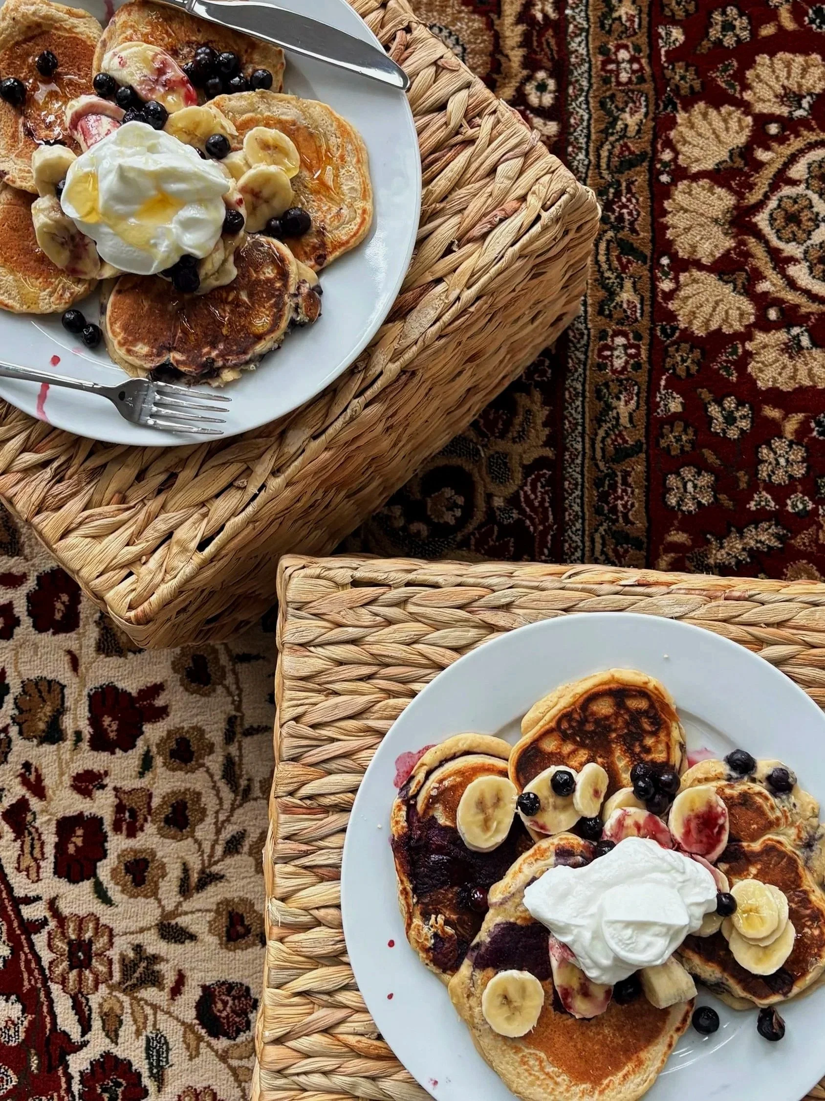 Plate of pancakes topped with bananas, blueberries, whipped cream, and syrup, on a wicker table with a Persian patterned rug underneath.
