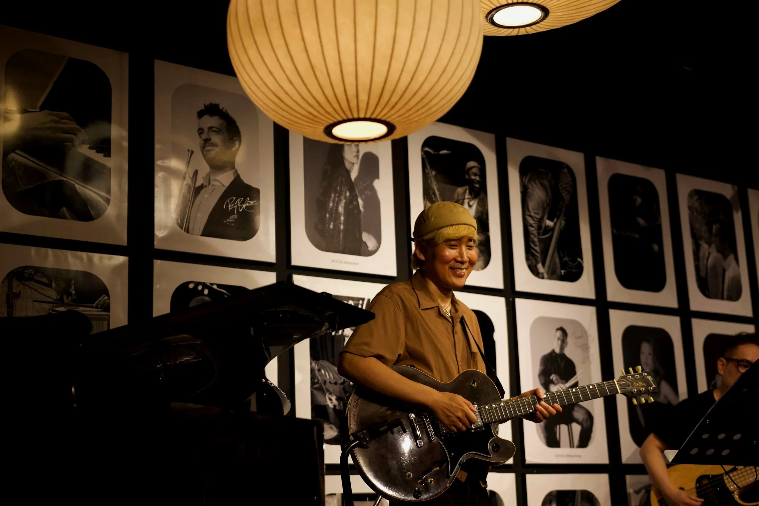 A musician with blonde hair and a brown shirt playing an electric guitar in a room with black and white photographs on the wall. Hanging from the ceiling is a large round paper lantern.