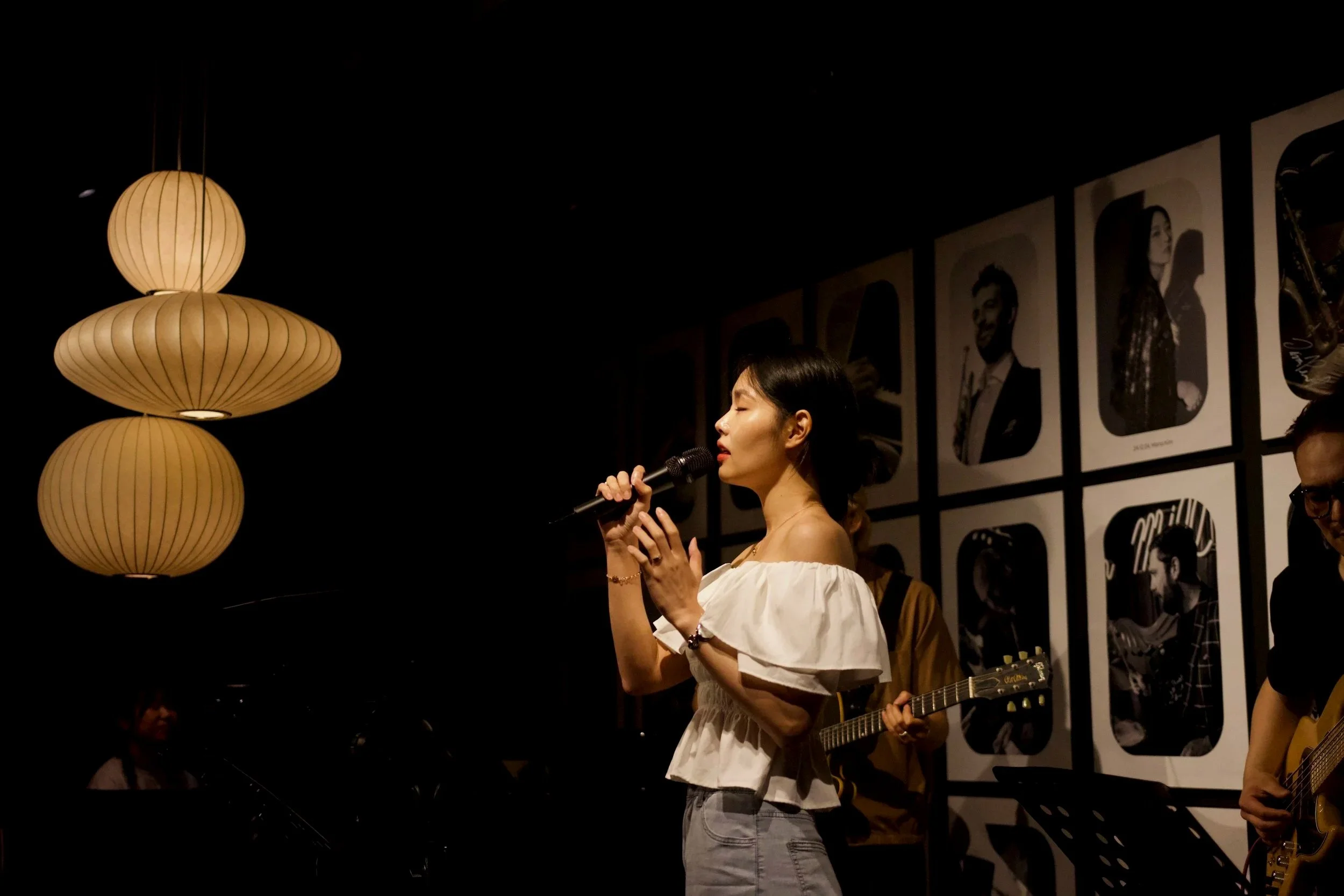 A woman singing into a microphone with eyes closed at a music event, surrounded by other musicians, with black and white portraits on the wall behind her and warm hanging lights to the left.
