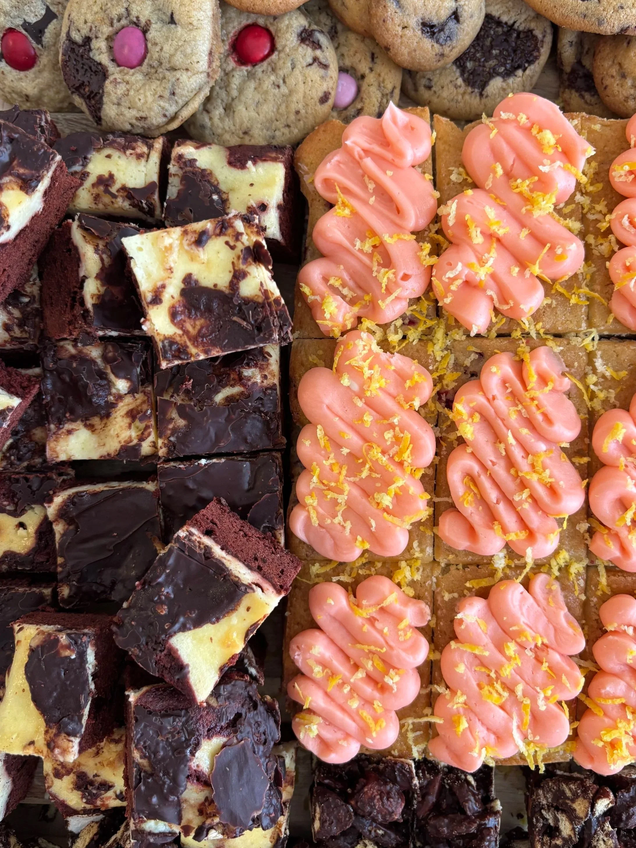 An assortment of baked goods including cookies, brownies, and frosted treats with pink icing and lemon zest.