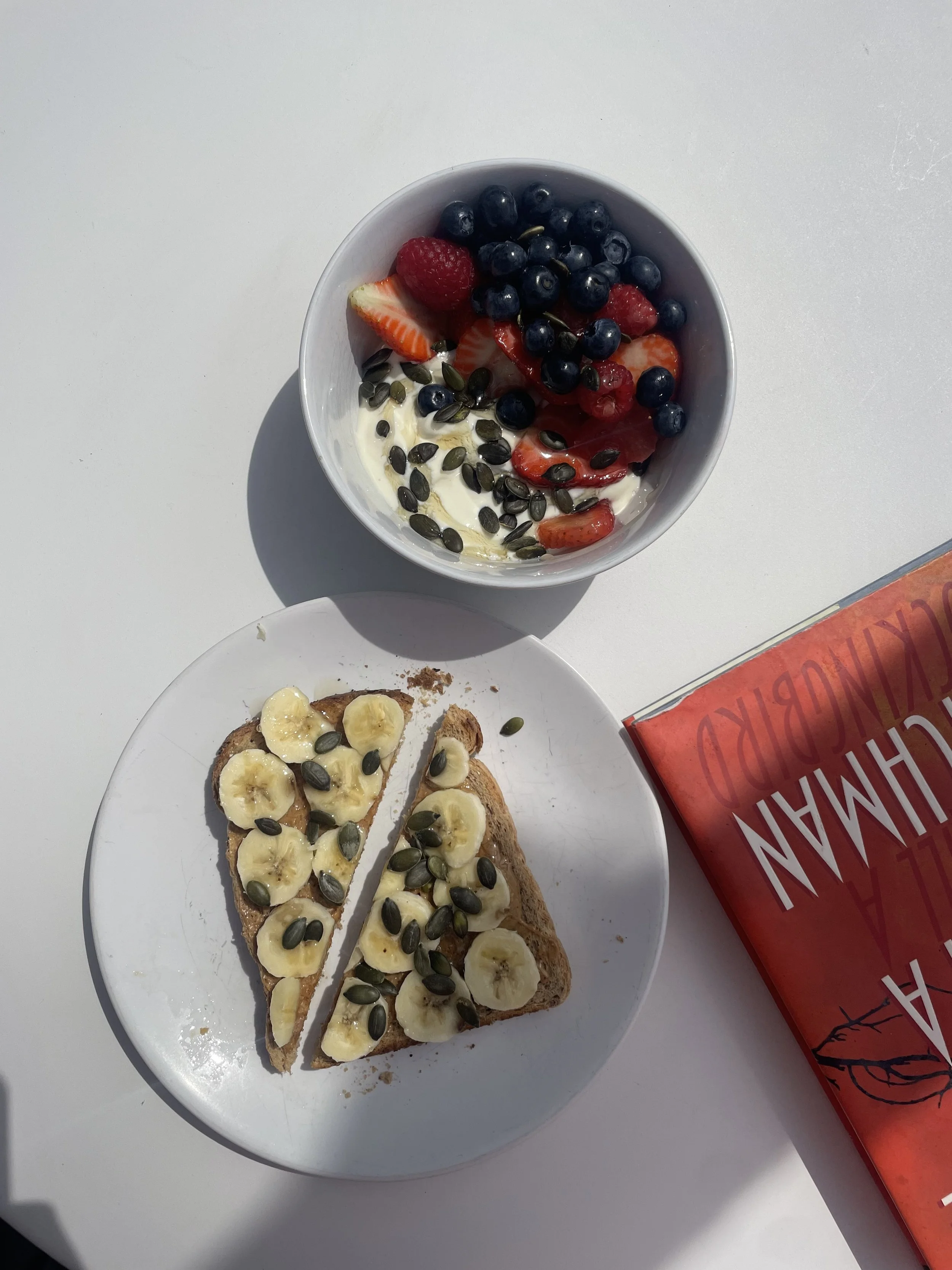 A white plate holding two slices of banana bread topped with banana slices and pumpkin seeds next to 'To Kill a Mockingbird'.