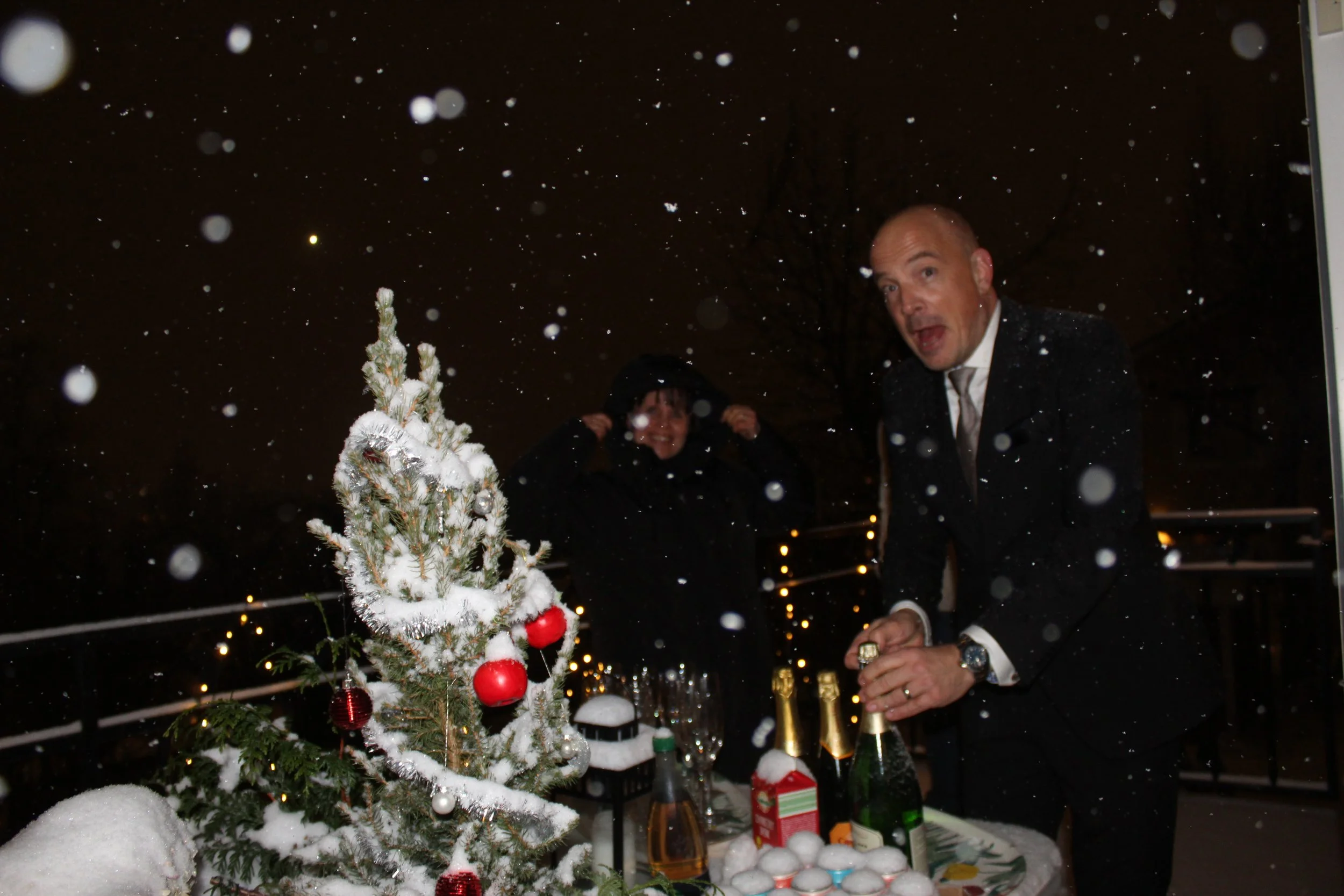 A small snow-covered Christmas tree decorated with red and white ornaments on a table outside at night, with two adults in winter clothing, one man dressed in a tuxedo, celebrating with champagne in the background