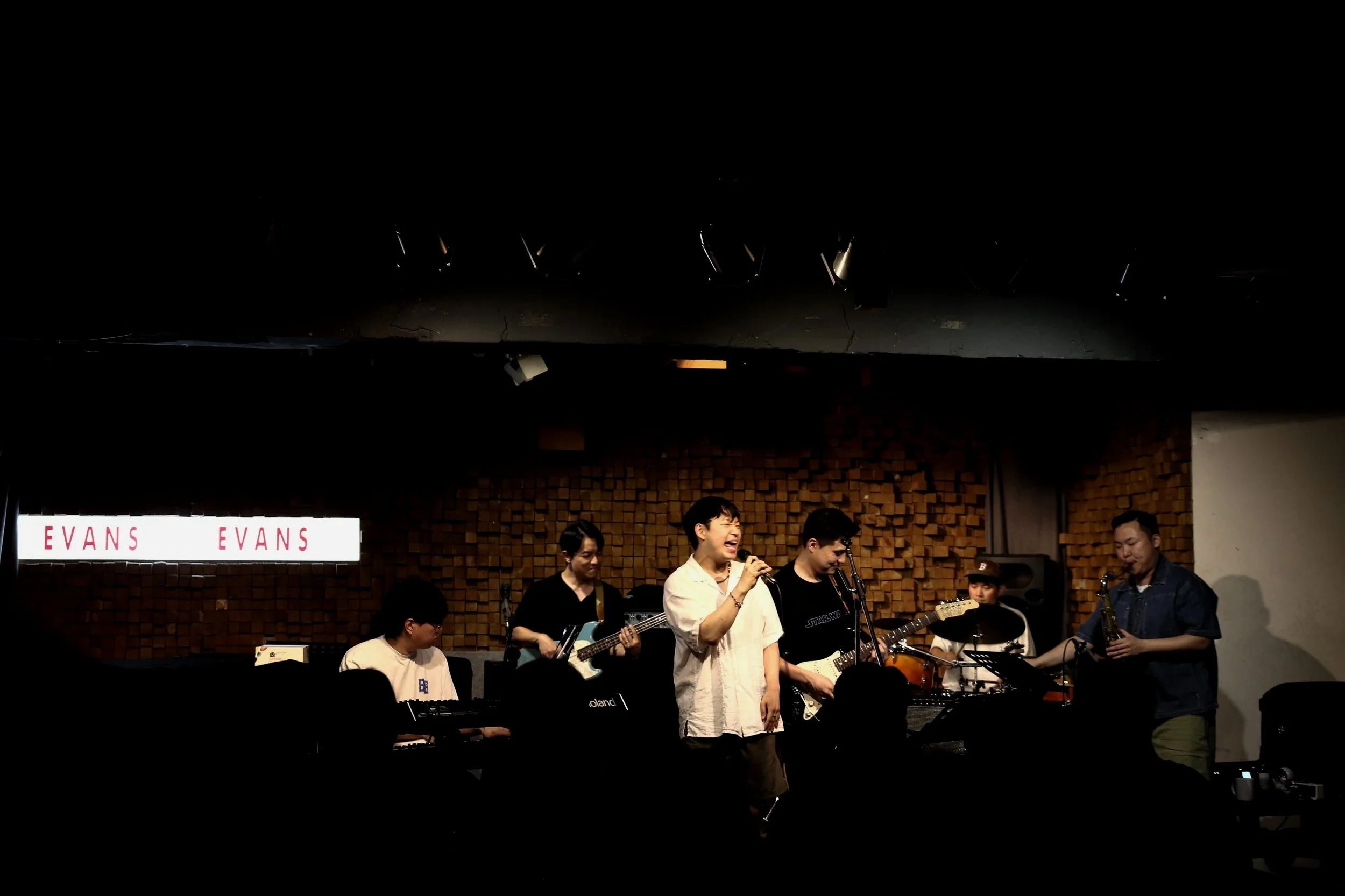 A group of musicians performing on stage with a brick wall background, including a singer, guitar players, and a saxophonist, with a sign that reads "EVANS" in the background.