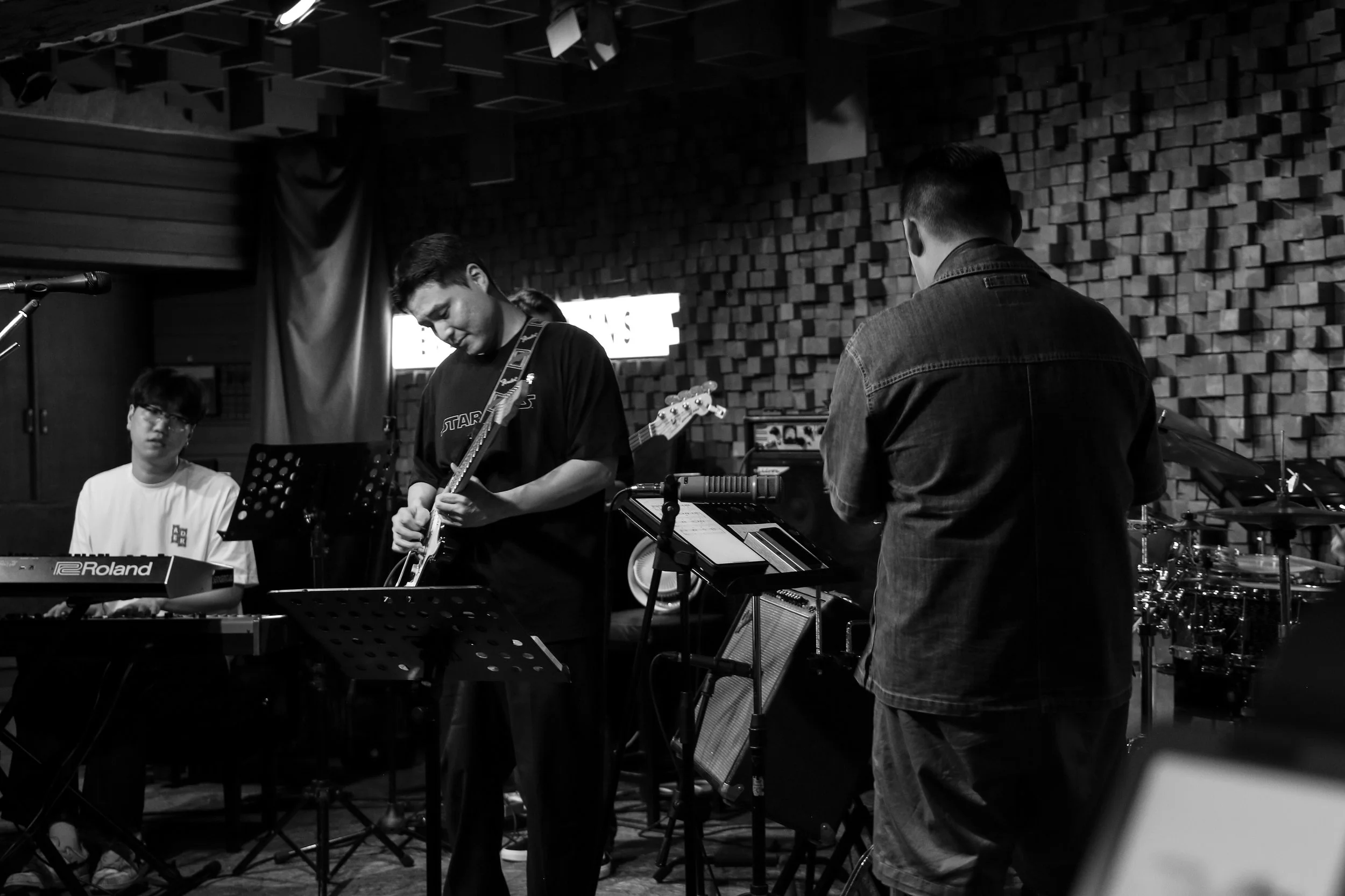 A black-and-white photo of jazz musicians on stage One is playing a keyboard, another is holding a guitar, and a third is working with electronic music equipment. They are in a room with acoustic foam panels on the wall.